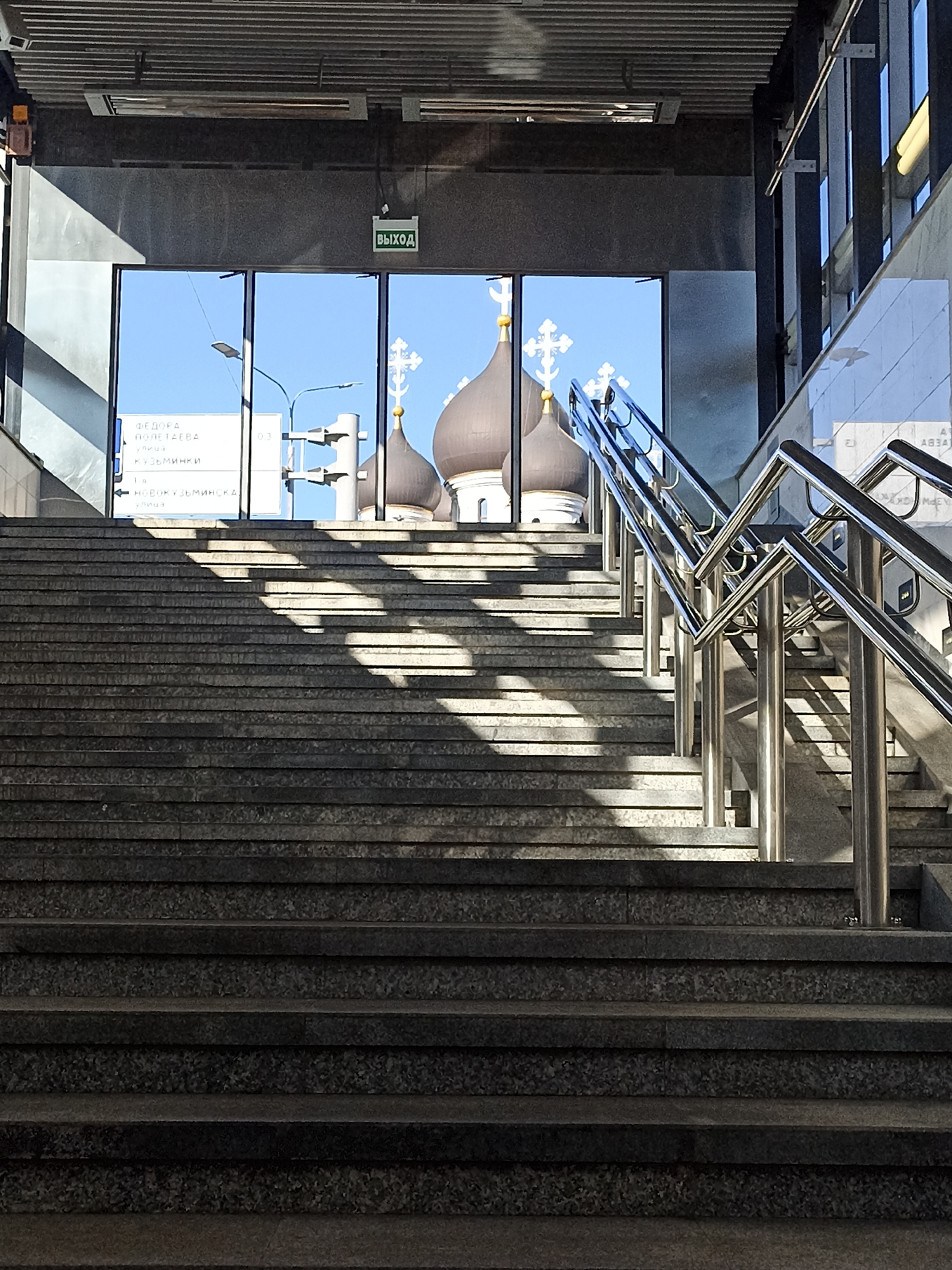 The image depicts an outdoor staircase leading up to a glass-enclosed walkway with a view of church domes and crosses in Moscow, Russia. The staircase is made of dark granite with distinct, horizontally textured steps. Sunlight casts sharp, diagonal shadows across the stairs. To the left, through the glass, a sign is visible with Cyrillic text, indicating street names such as "ФЁДОРА ПОНЕТАЕВА" (Fyodor Ponetaev) and "КУЗЬМИНКИ" (Kuzminki). The walkway features a prominent green "ВЫХОД" (Exit) sign. On the right side of the staircase, a modern stainless steel handrail with multiple angled sections guides the ascent. The overall environment suggests an urban setting, possibly near a transit hub or a public building, with a blend of architectural styles.