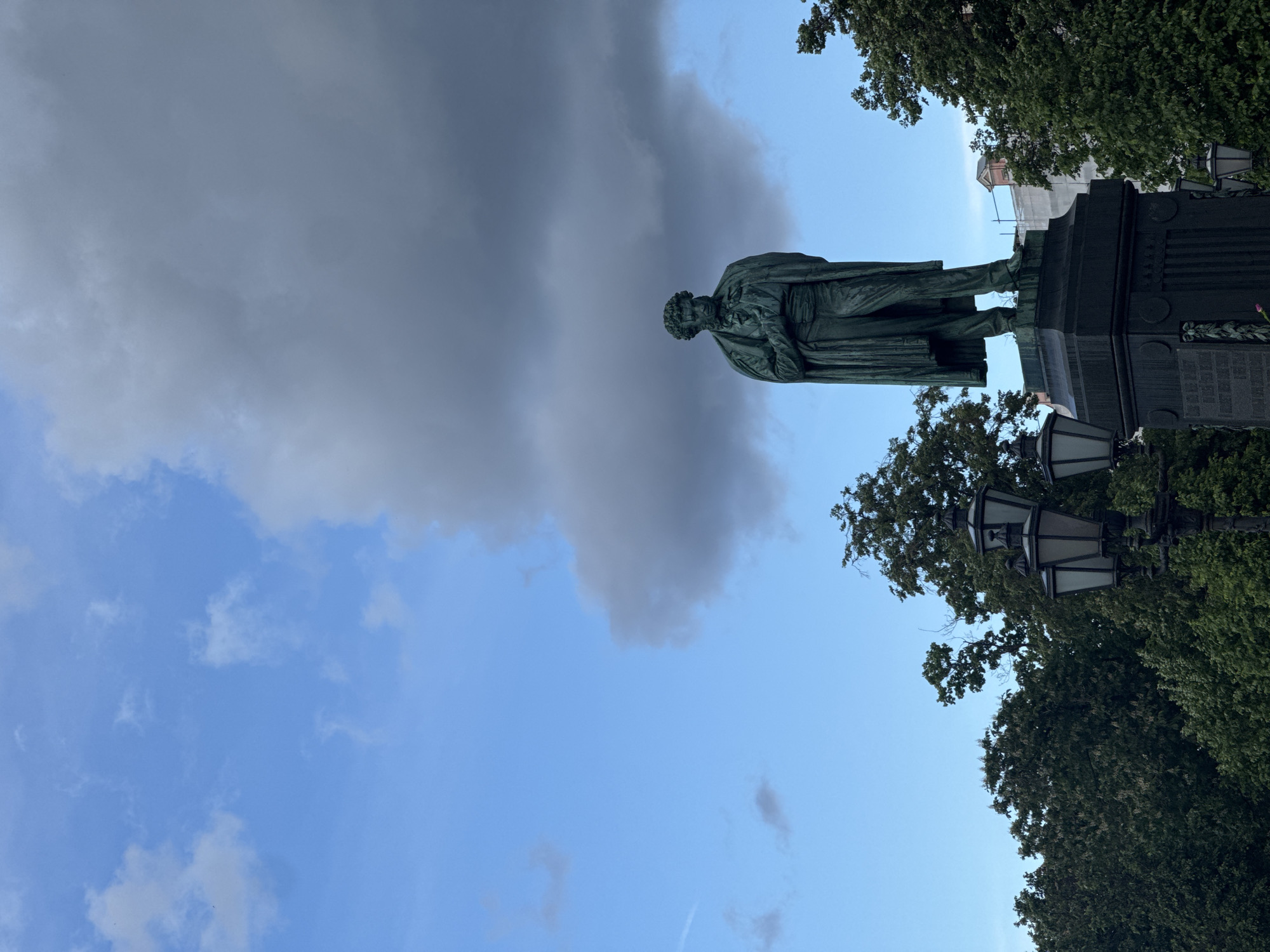 The image shows the Monument to Alexander Pushkin on Pushkin Square in Moscow, Russia. The bronze statue of the poet stands on a tall, dark pedestal adorned with engravings and surrounded by foliage and old-fashioned street lamps. The statue is viewed from below, the subject appears to be standing against a backdrop of blue sky and imposing grey clouds.