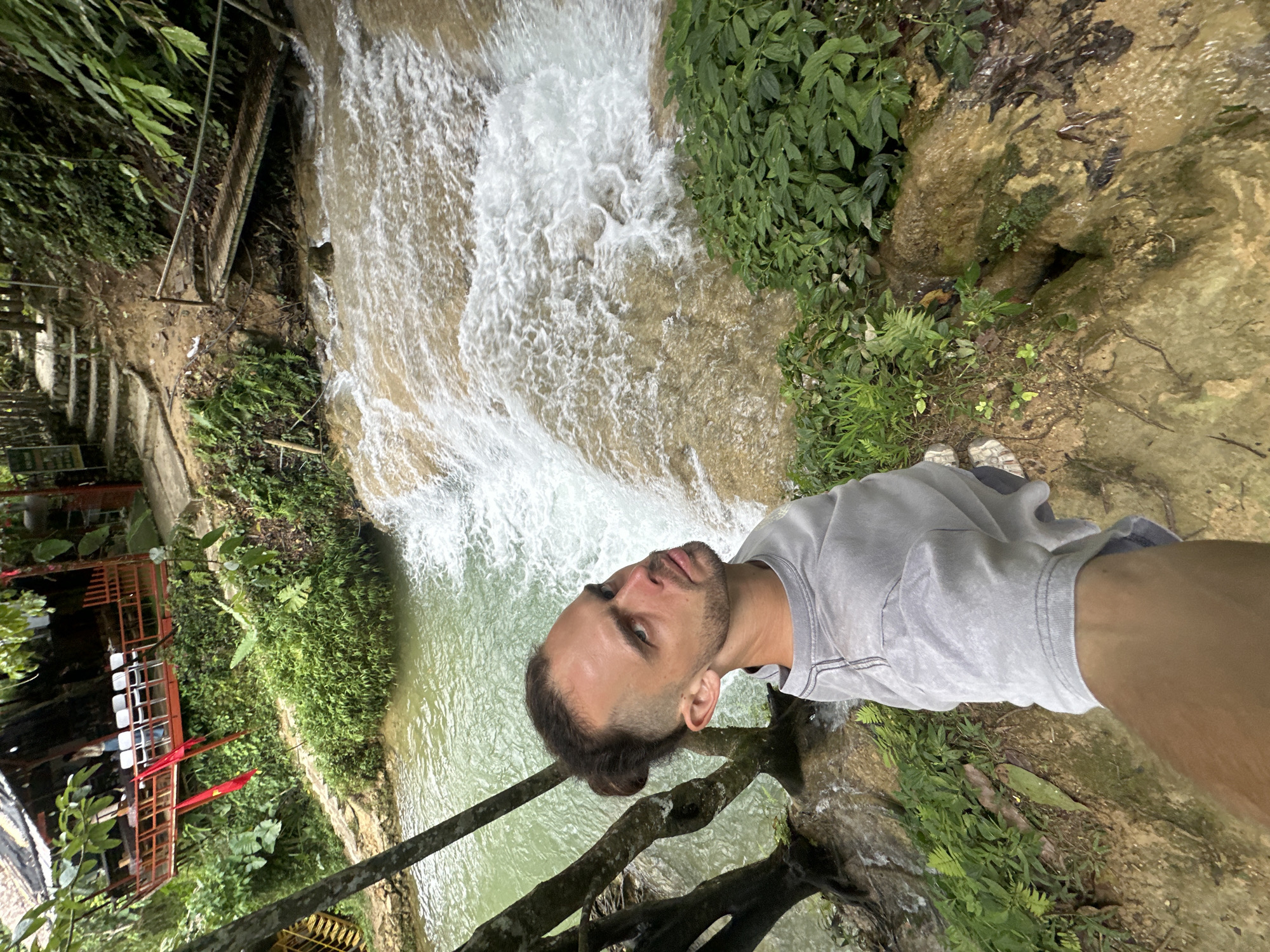 A man with his hair pulled back into a bun is taking a selfie in front of a waterfall in Cổ Lũng Commune, Vietnam. He is wearing a light grey t-shirt and white sneakers. The waterfall cascades over beige rocks into a pool of turquoise water. Lush green vegetation surrounds the waterfall, and in the background, a wooden structure with red railings and white chairs is visible, with a red flag hanging from it. A pathway with stone steps leads up the hill behind the structure.