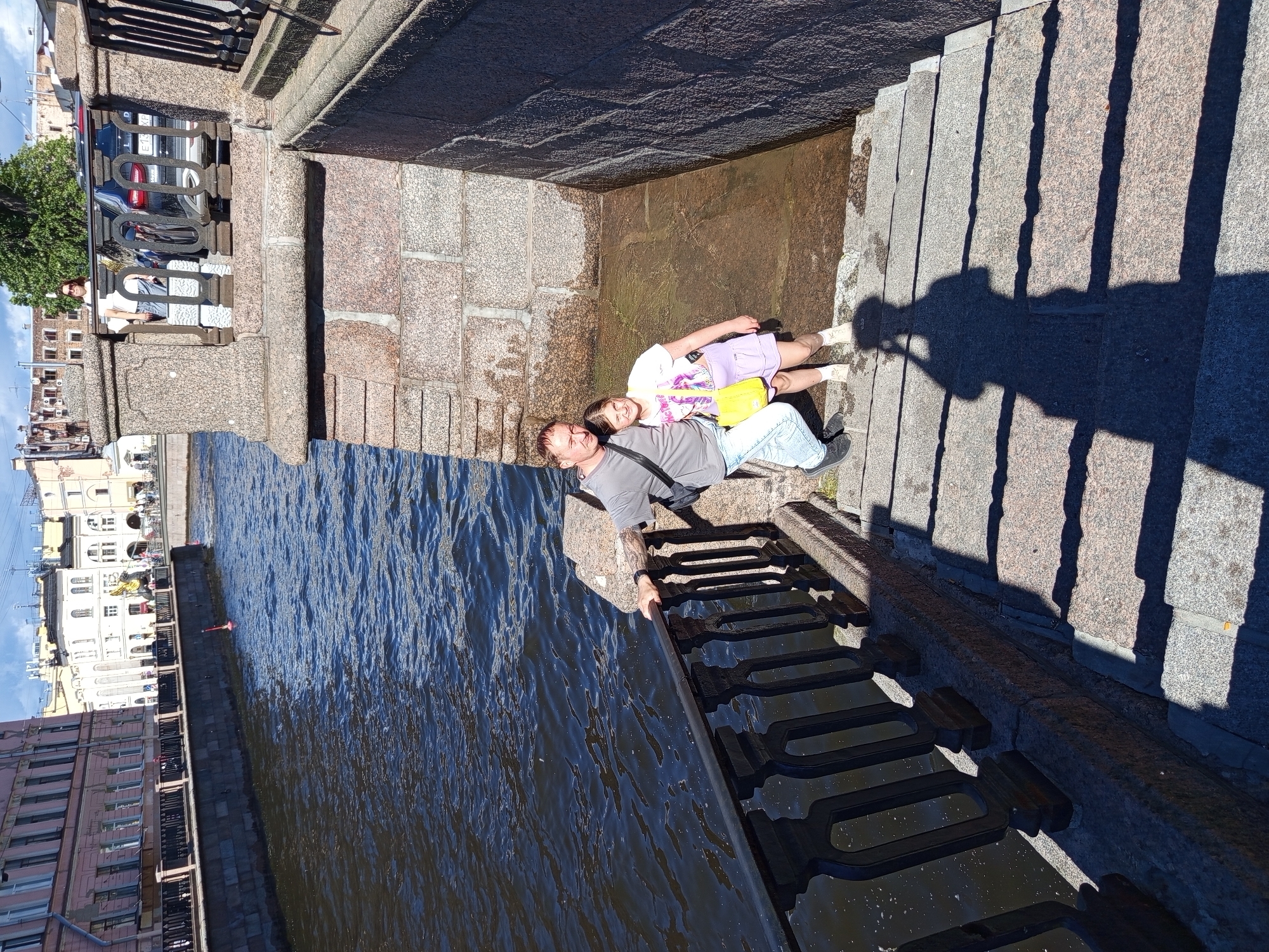 In Saint Petersburg, Russia, a man and a young girl are posing for a photograph on stone steps descending towards a canal. The man, wearing a grey t-shirt and light blue jeans, has his arm around the girl. The girl, in a white t-shirt and a light purple skirt, carries a yellow bag. They are positioned on a wide, granite staircase. A dark metal railing runs along the canal side. The canal water is dark blue and reflects sunlight. Buildings line the opposite bank of the canal. The overall scene suggests a sunny day at a historic urban location.