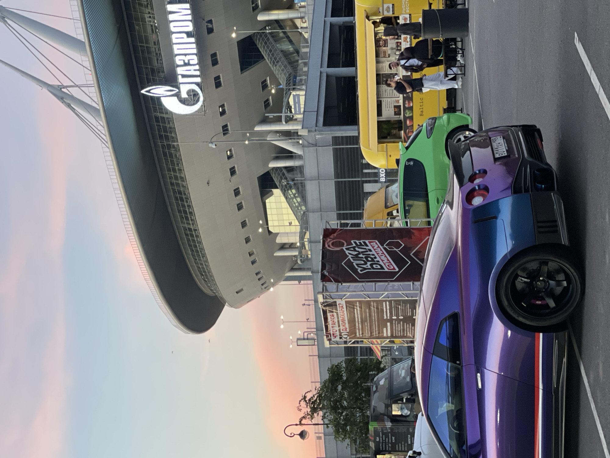 In an outdoor setting, a purple and blue Nissan GT-R is parked in the foreground, facing away from the camera. The car has a spoiler and custom wheels. Behind the car, a bright yellow food truck is visible with people standing and interacting near it. A green compact car is parked in front of the food truck. In the background, a large modern stadium with a prominent "GAZPROM" logo is illuminated against a twilight sky with pink and blue hues. Several lampposts and trees are also present in the scene. Two banners stand between the car and the food truck; one has text in Russian and the other has the words "YUKA DRIVE GYMKHANA". The ground is paved with asphalt.