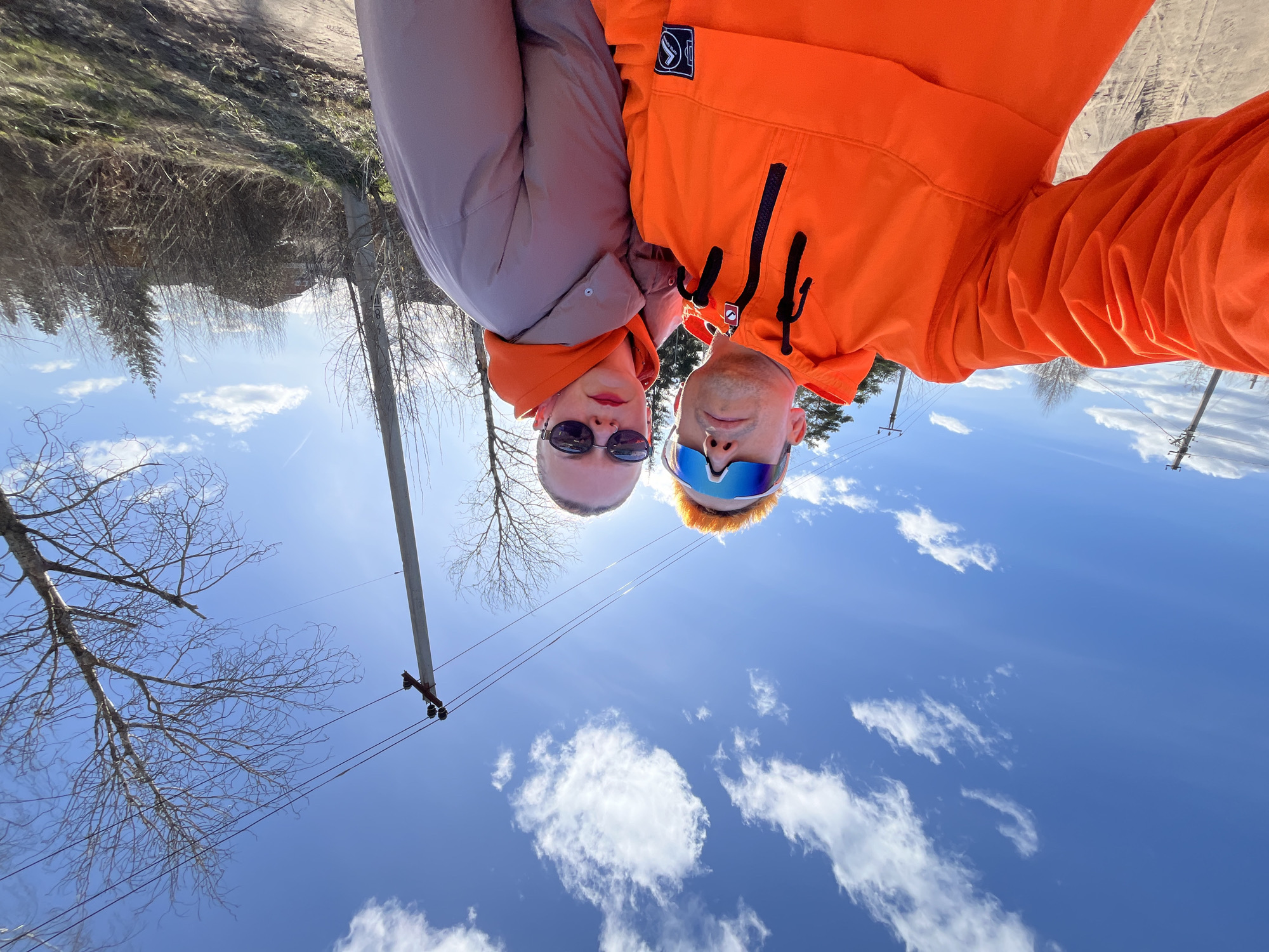 The image is taken in Chaikovsky, Russia, and shows two people taking a selfie. The people are a man and a woman. The man is wearing an orange hooded jacket and reflective sunglasses. The woman is wearing sunglasses and a light gray jacket. They appear to be standing outside on a sunny day with a blue sky and white clouds. Bare trees are visible in the background along with a utility pole.