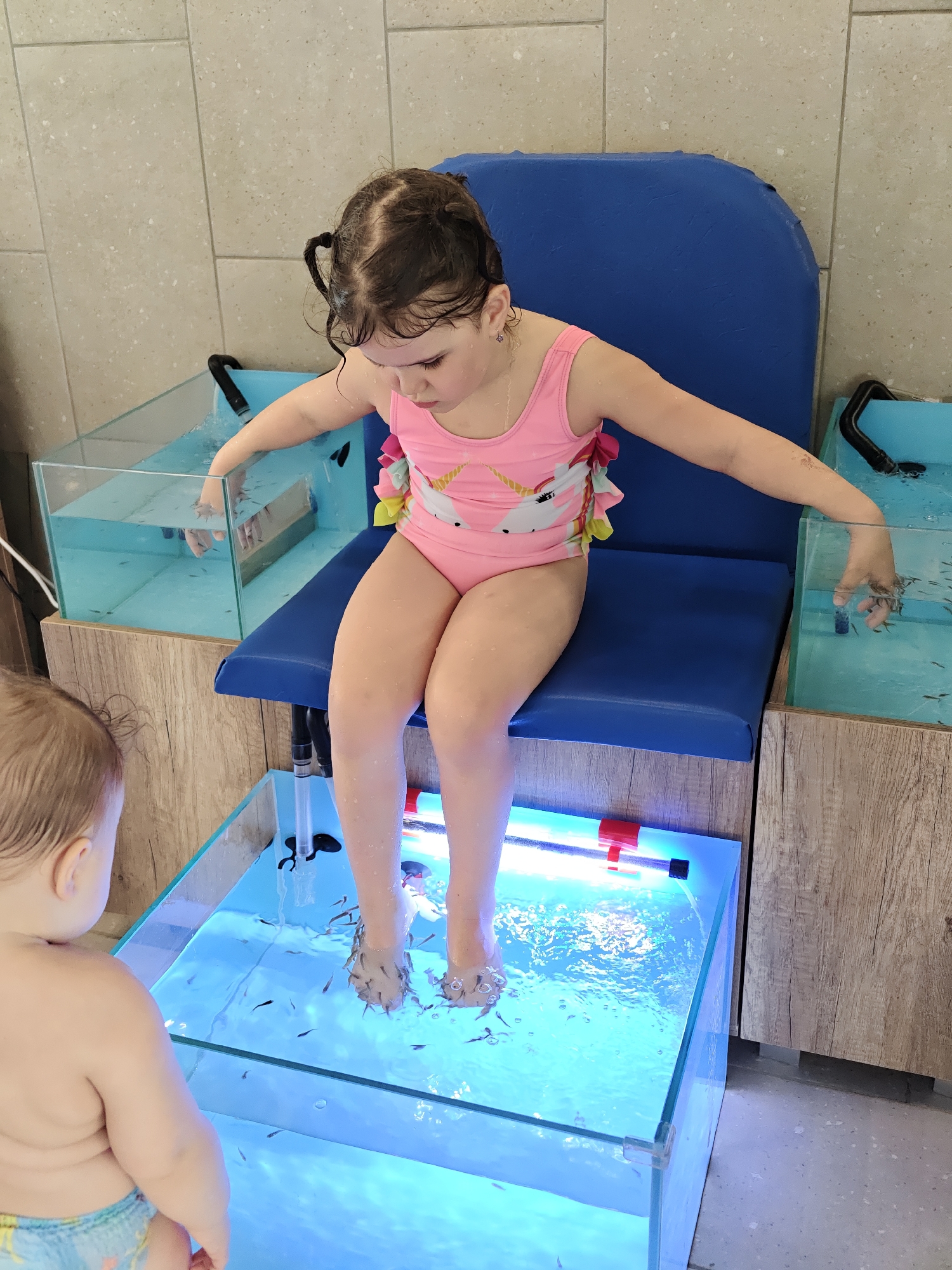 A young girl with dark hair styled in pigtails, wearing a pink swimsuit featuring a unicorn design, is seated on a blue padded chair. She is participating in a fish spa treatment, with her feet submerged in a glass tank filled with water and numerous small Garra rufa fish. Her hands are also placed in two separate, smaller glass tanks, one on each side, which also contain fish. The tanks are illuminated with a blue light, and various pipes and filtration elements are visible, including a red and black fixture in the main foot tank.
The girl is looking down intently at the fish, displaying an expression of curiosity and concentration. To the left of the frame, partially visible from behind, is a baby or toddler with light hair, wearing a diaper or swim trunk, appearing to observe the scene.
The setting is an indoor space with light beige/grey tiled walls and wooden paneling surrounding the fish tanks. The overall environment suggests a commercial fish spa or a similar establishment offering this type of treatment, located in Votkinsk, Russia. No visible text is present in the image.