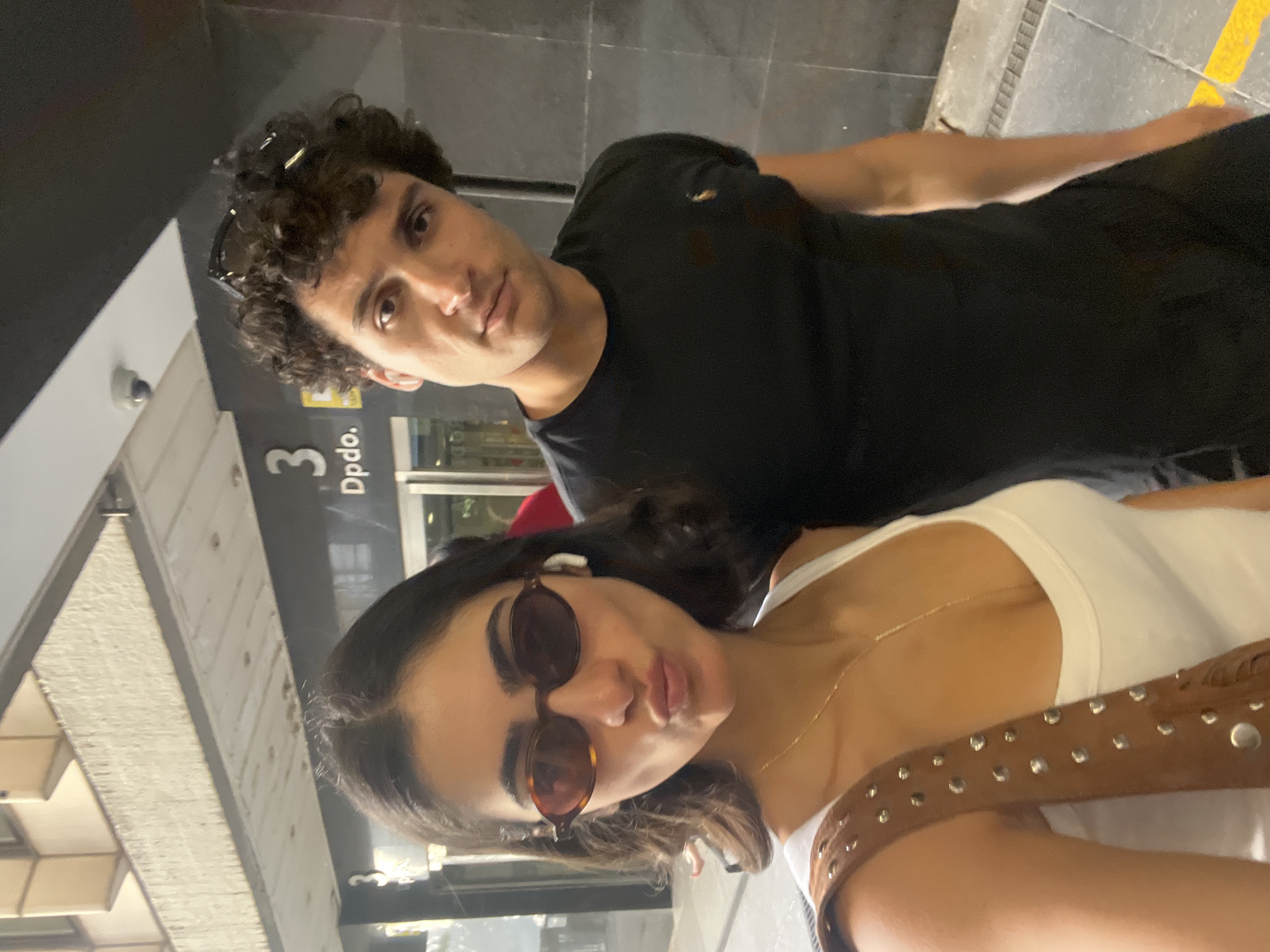 Two people are standing close to each other in what appears to be an urban environment, likely a subway or train station. A man with curly hair is wearing a black T-shirt and sunglasses. The woman has sunglasses and is wearing a white tank top. The number "3" is visible on a sign along with "Dpdo". There's a security camera and what appears to be an outdoor view visible through a glass door. The scene is possibly in Madrid, Spain.