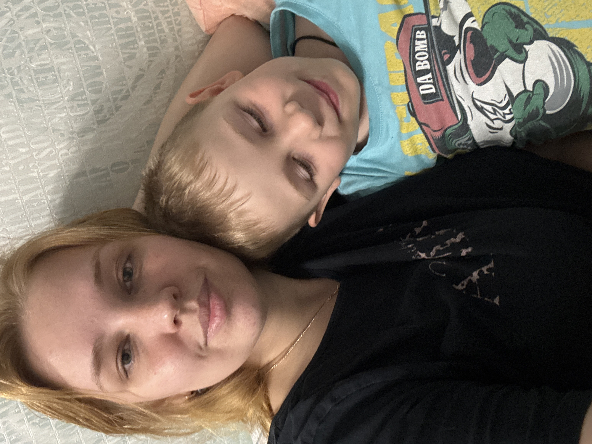 The image shows a woman and a young boy lying on a bed, possibly in a home environment. The woman has light skin, blonde hair, and is wearing a black shirt with a necklace. The boy has light skin, blonde hair, and is wearing a light blue t-shirt with a cartoon graphic labeled "DA BOMB." They are lying close together, possibly embracing or resting. The bed has a patterned white bedspread. The location in the city of Rusanovo, Russia, is not confirmed and is not determinable from the image alone.