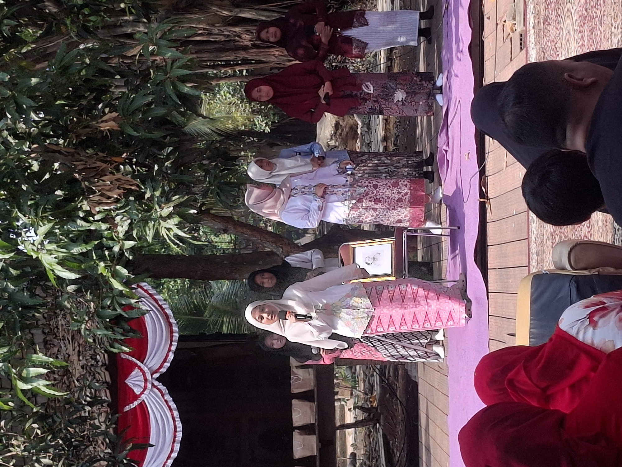 In the city of South Tangerang, Indonesia, an outdoor stage with a red and white draped backdrop is set up beneath lush trees. A young woman wearing a white hijab and a white patterned top with a pink patterned skirt stands at a microphone, addressing an audience. Behind her, several other women in hijabs and traditional attire are visible, some holding microphones and others standing or sitting. A framed portrait of an older individual rests on a chair near the speaker. The stage is covered with a purple cloth, and the audience is partially visible in the foreground, with some individuals wearing red hijabs. The overall scene suggests a cultural or religious event taking place outdoors.