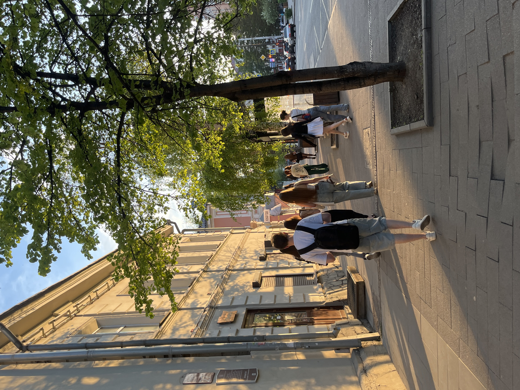 The image depicts a group of people walking along a sidewalk in Krasnoyarsk, Russia. The sidewalk is made of rectangular pavers, and a row of trees lines the street, casting shadows on the ground. A light-colored building stands adjacent to the sidewalk. The people are walking in the same direction, their postures and gaits indicative of walking. The scene appears to be bathed in sunlight, suggesting it is daytime. Several plaques are visible on the building wall, potentially providing historical or informative context. A road with car traffic is visible further into the shot.