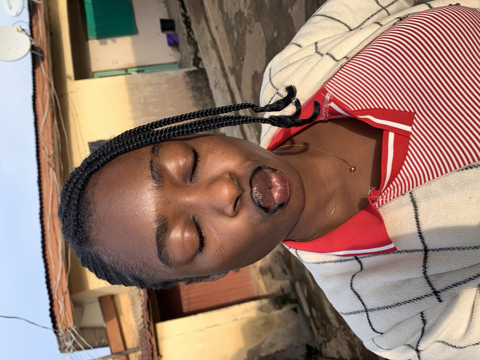 A medium shot photograph shows a young Black woman with her eyes closed and her lips puckered, as if blowing a kiss. She has braided hair, with two braids falling over her shoulder. She is wearing a white jacket with black lines, and a red and white striped shirt. The background shows a building with a brown roof, and two satellite dishes. The scene appears to be outdoors, possibly in a residential area. The lighting suggests it is either early morning or late afternoon. The location is identified as Rafin Par, Nigeria.