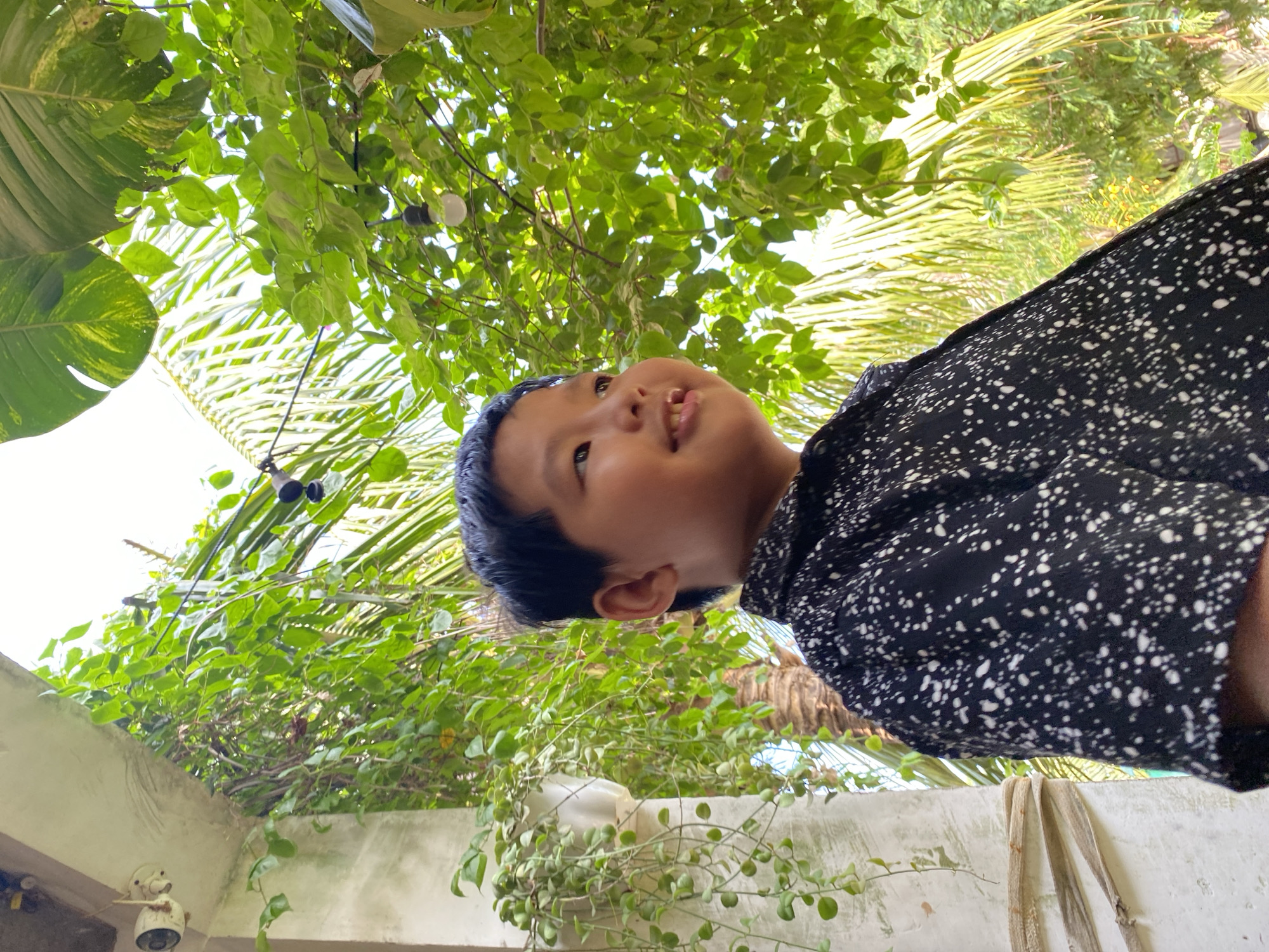 An outdoor, low-angle shot shows a young East Asian boy looking to his right and smiling, with his teeth visible. He is wearing a black shirt with white speckles. He is positioned in the foreground, with lush green foliage and palm fronds filling the background. Sunlight filters through the leaves, creating bright highlights. To the left of the boy, part of a white, textured wall is visible, with a hanging plant and a security camera mounted on an overhang. The overall scene is bright and airy, suggesting a warm, tropical or subtropical environment.