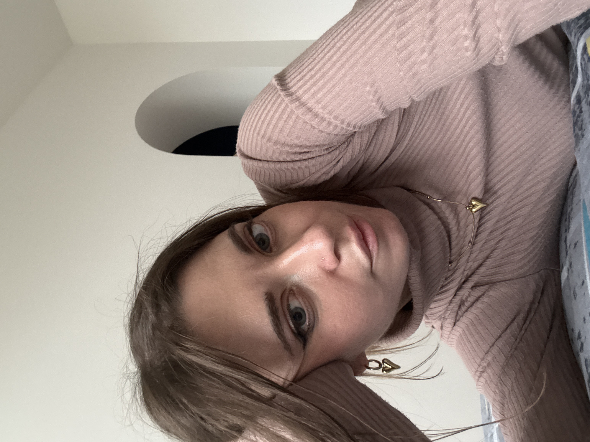 A woman is seen in a close-up shot, with her face angled towards the camera. She has light brown hair and fair skin. She is wearing a pink turtleneck sweater and gold earrings. A necklace with a small heart pendant is visible. A portion of a white wall with a rounded architectural element can be seen in the background. There is no indication of the location being Moscow, Russia in the image.