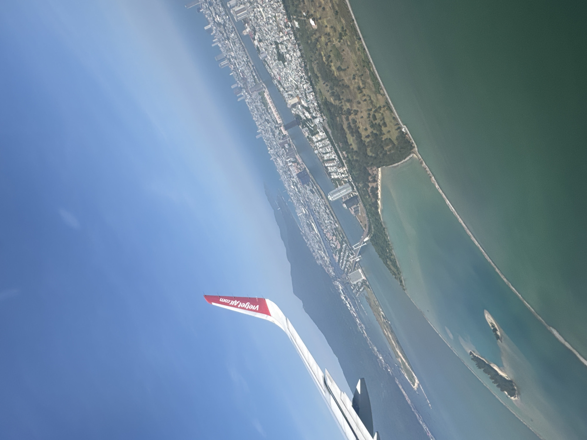The image shows a view from an airplane, presumably a Vietjet Air flight based on the visible wing. Below, the urban sprawl of Đà Nẵng, Vietnam, is visible, bordered by a coastline and the sea. The city's buildings are densely packed. A river or waterway cuts through the city. Beyond the city, a long, narrow stretch of land covered in greenery parallels the coastline. Further out, islands are visible in the water. The sky is clear and blue.