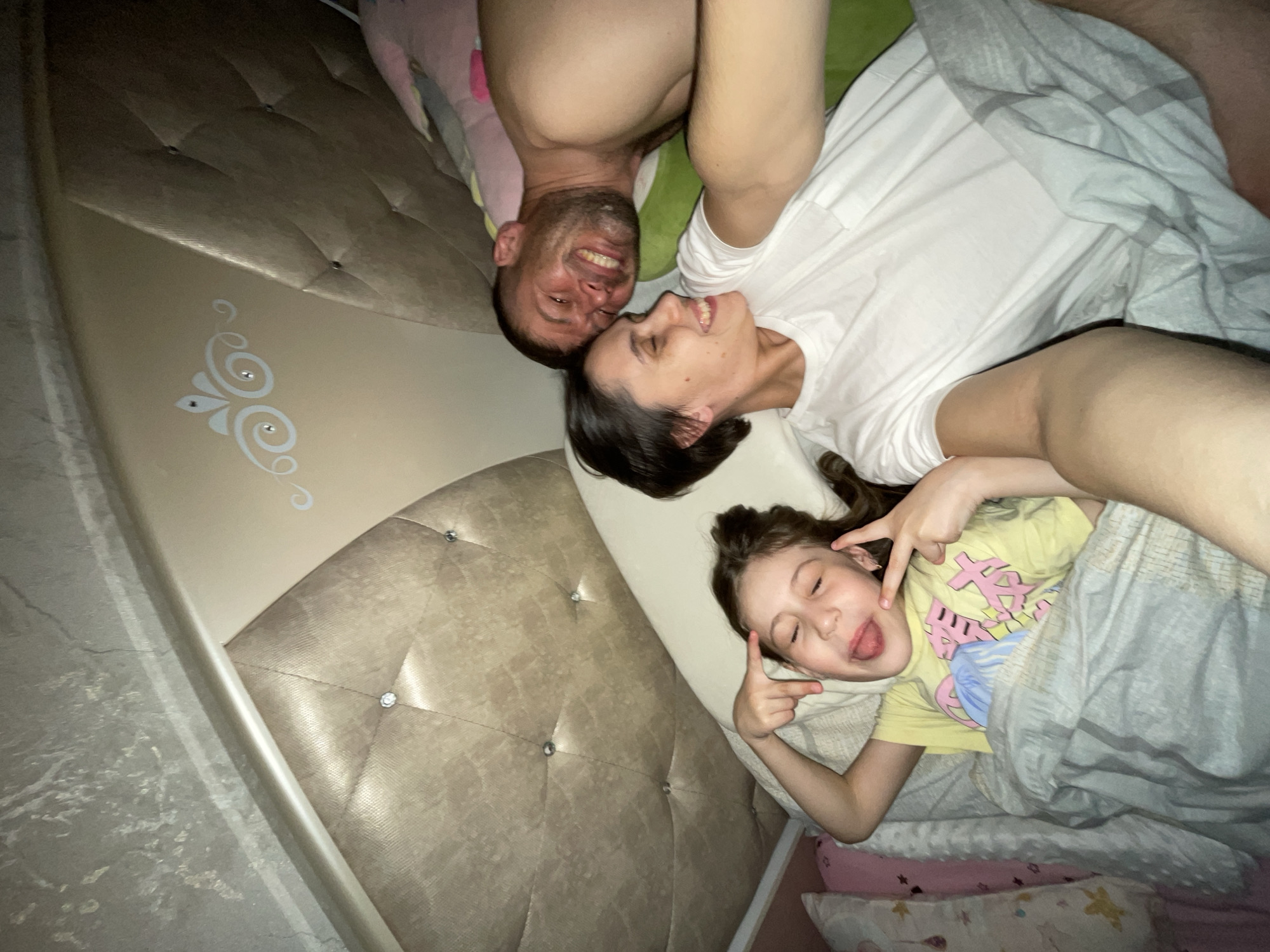 A family of three poses in bed for a selfie indoors. An adult male with short dark hair and a trimmed beard is at the top of the frame, upside down, smiling widely. Beneath him is an adult female with dark hair, also smiling and looking upwards towards the man. At the bottom of the frame is a young girl lying in bed, making a silly face with her tongue out and her eyes closed, also making a peace sign with her fingers. They appear to be in a bedroom, with a light-colored, quilted headboard visible behind them. The room has beige tones and is dimly lit. The location is Izhevsk, Russia.