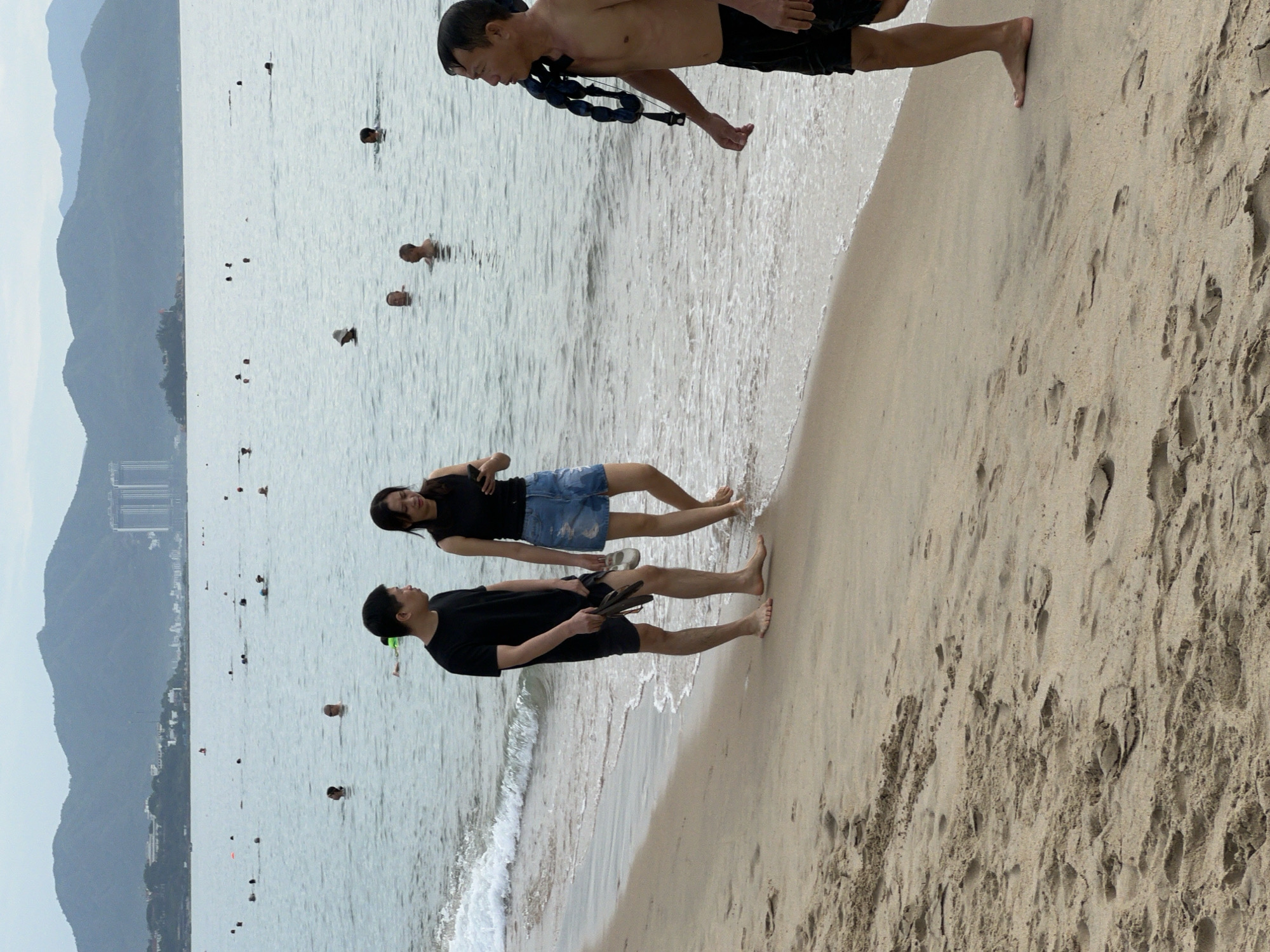 A group of people are seen on a sandy beach in Nha Trang, Vietnam, with the sea and a hilly coastline in the background. In the foreground, a man in black shorts walks away from the camera, carrying a blue life vest over his shoulder. To his left, a man and a woman are walking along the shoreline. The man is wearing a black t-shirt and shorts, holding flip-flops in his hand. The woman is in a black sleeveless top and denim shorts, looking at her phone. Numerous people are visible in the water, some swimming and others wading. The sea is calm with small waves reaching the shore. The sky is overcast. The sand on the beach is textured with footprints and small sand formations.