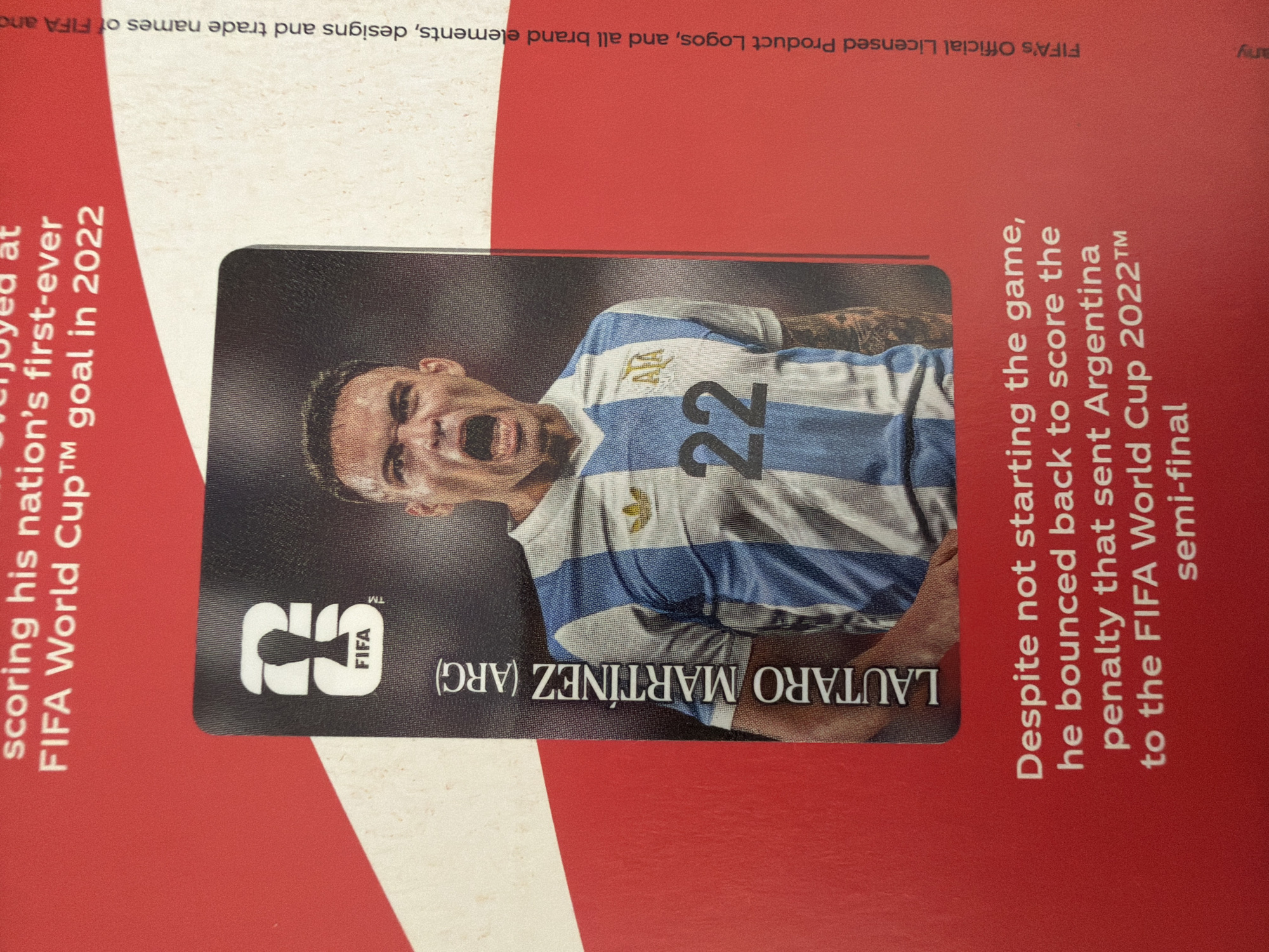The image shows a close-up of a trading card featuring a male soccer player wearing an Argentina national team jersey with the number 22. The player has dark hair and is shown with his mouth open, appearing to be shouting. The jersey is blue and white striped. The card has a FIFA logo at the top and the player's name, "LAUTARO MARTÍNEZ (ARG)", is written vertically along the left side. The card is placed on a red surface with white stripes. Text in white is visible on the red surface, describing the player's achievements in the FIFA World Cup 2022. Vertical text on the right side reads "FIFA's Official Licensed Product Logos, and all brand elements, designs and trade names of FIFA and". The image does not contain any information or context related to the city of Zlatitrap, Bulgaria.