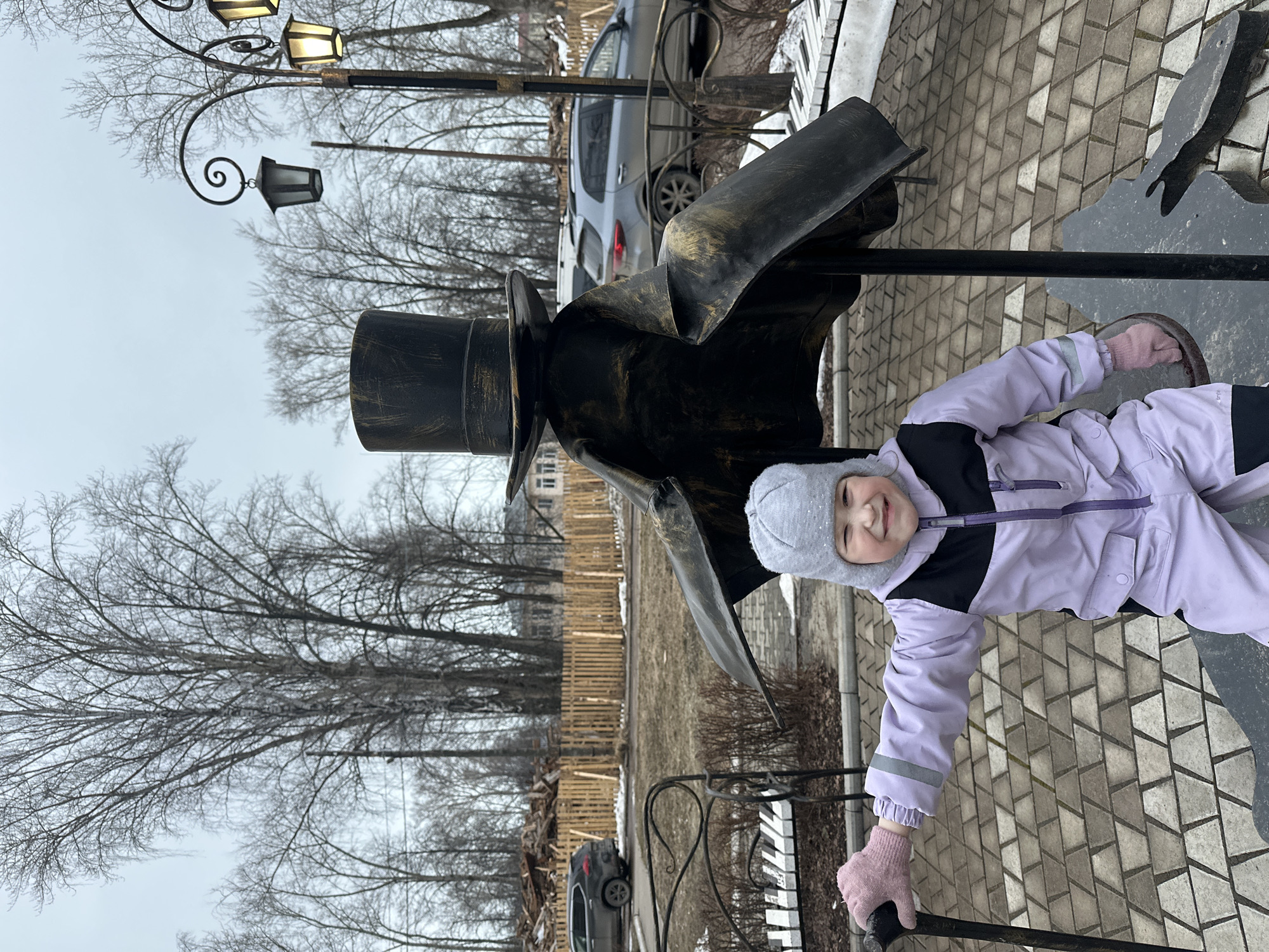 A young girl, wearing a light purple snowsuit with black accents and a gray knit hat and pink mittens, is positioned on a sidewalk in Chaikovsky, Russia. She is smiling and appears to be holding onto a decorative, dark metal sculpture of a figure wearing a top hat and cloak. The sidewalk is paved with light gray patterned bricks. In the background, there are bare trees, a light-colored wooden fence, and parked cars. A decorative lamp post is visible in the upper left corner. The sky is overcast and gray. The sculpture and the girl appear to be interacting as though they are friends.