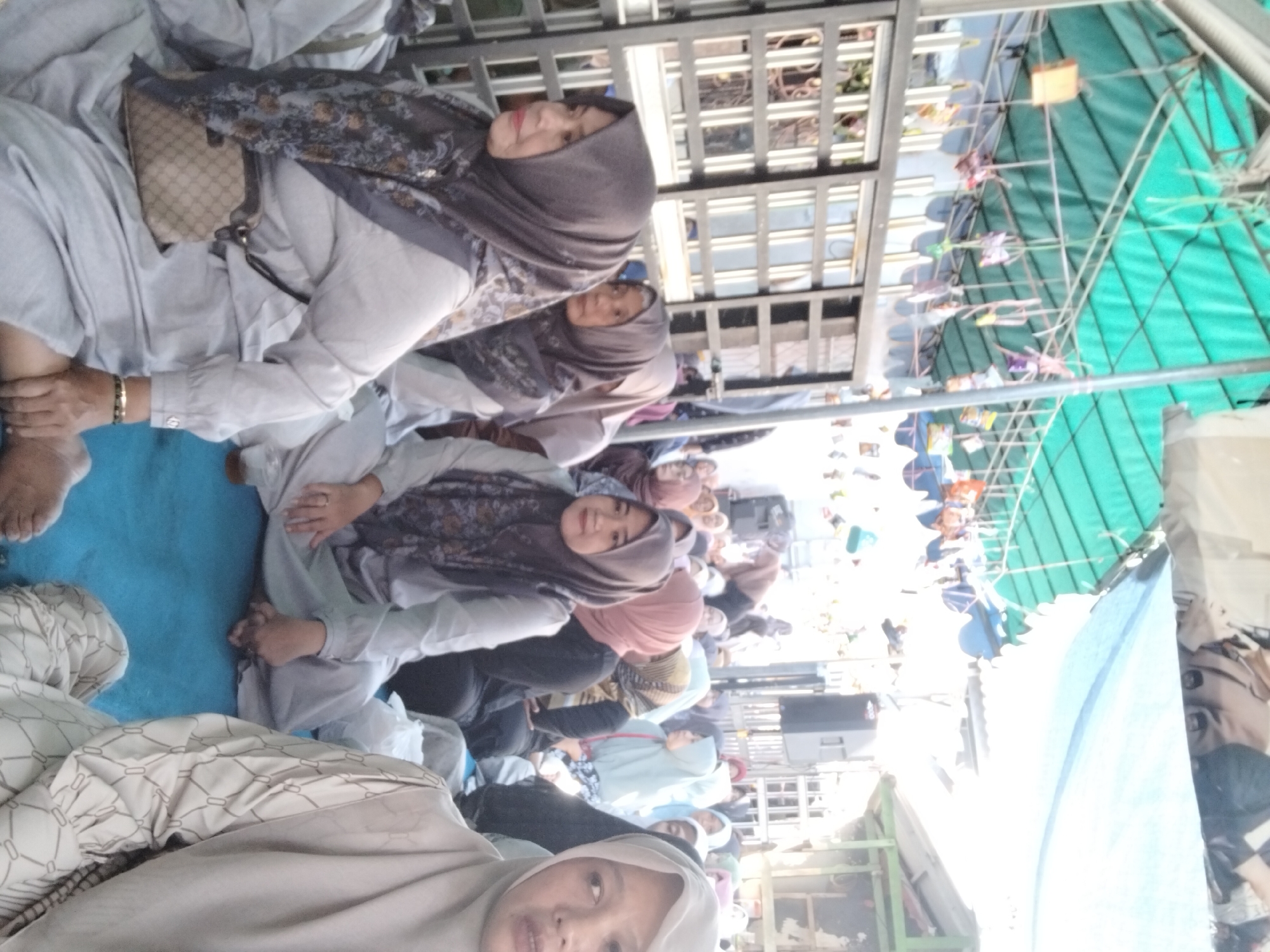 The image captures an outdoor gathering in Kepuh Kiriman, Indonesia, likely during a daytime event. A group of individuals, predominantly women, are seated on a blue mat under a green tarp. They are all wearing headscarves, with many also wearing long, loose-fitting clothing in muted colors. Some women are looking to the side, while others appear to be interacting with each other or observing something out of frame. In the background, there are structures that suggest a marketplace or community area, with various stalls and some metal fencing. Colorful decorations, possibly bags or streamers, are hanging from a structure above the tarp. The overall atmosphere appears to be one of community and social engagement.