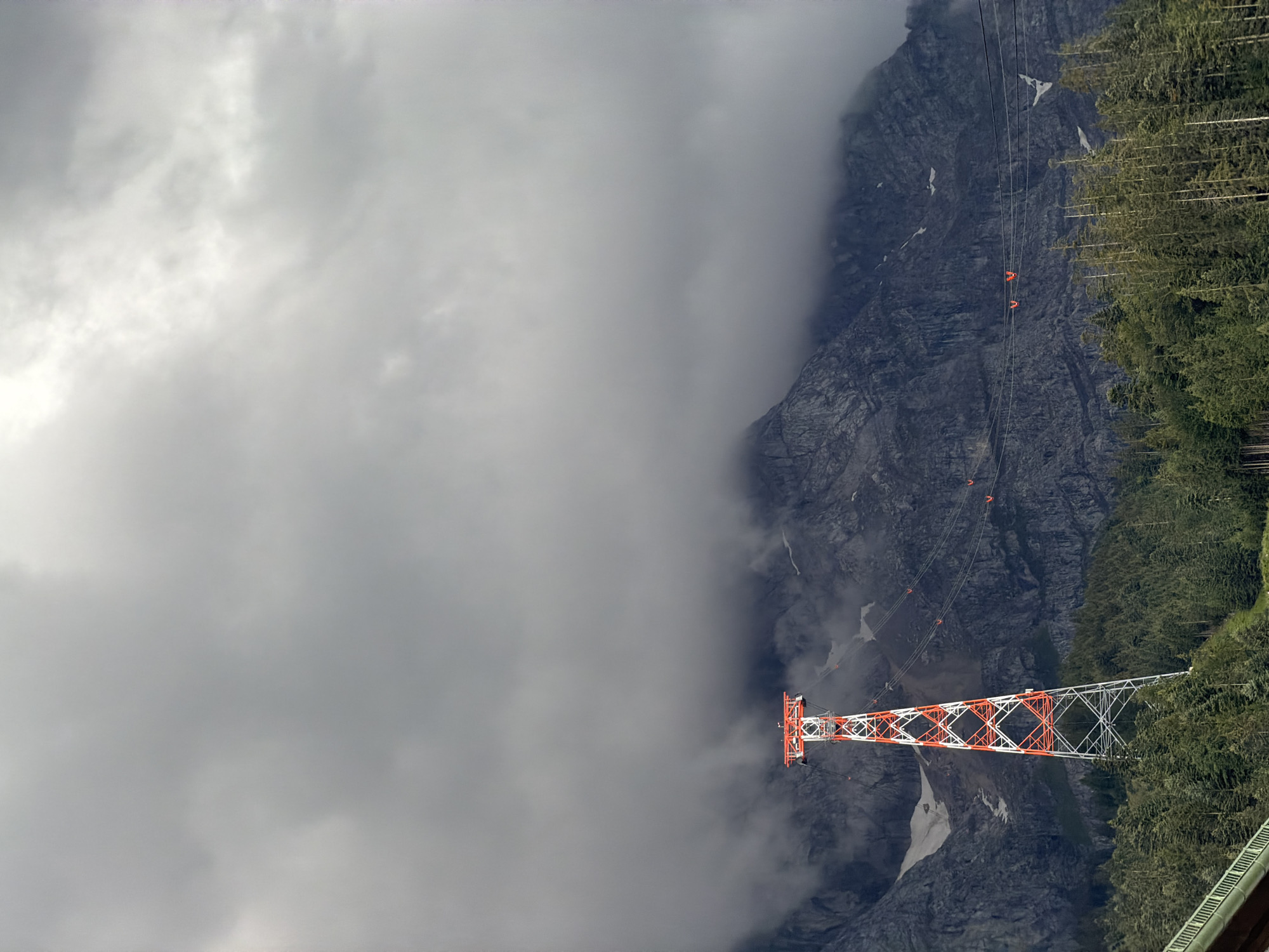 The image shows a tall, red and white lattice tower, likely a support for a cable car or ski lift, situated on a wooded mountainside. The tower is located in Grainau, Germany, at the base of the Zugspitze mountain. The sky above is overcast with dense grey clouds that partially obscure the rugged mountain peaks in the background. Several small, orange cable cars are visible suspended on cables stretching across the scene, leading towards the obscured mountains. The foreground features a dense forest of evergreen trees. A portion of a railing or bridge structure is visible in the lower left corner of the image.