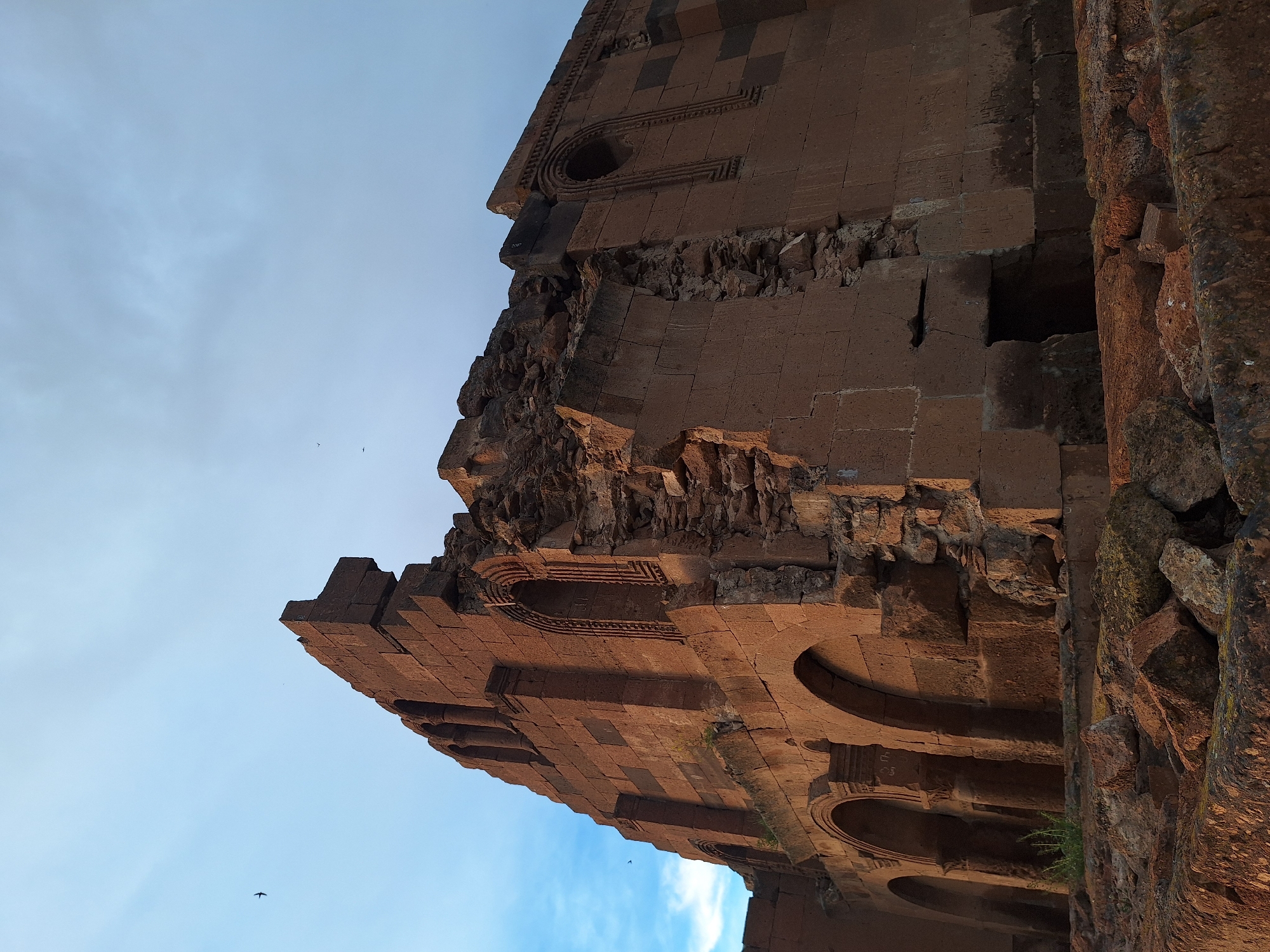 The image shows a portion of a damaged stone building, possibly a church or historical structure, located in Anipemza, Armenia. The architecture features brick or stone construction with arched windows or openings. There is visible deterioration and collapse of the structure, with fragmented walls and debris. The sky is partly visible in the background.