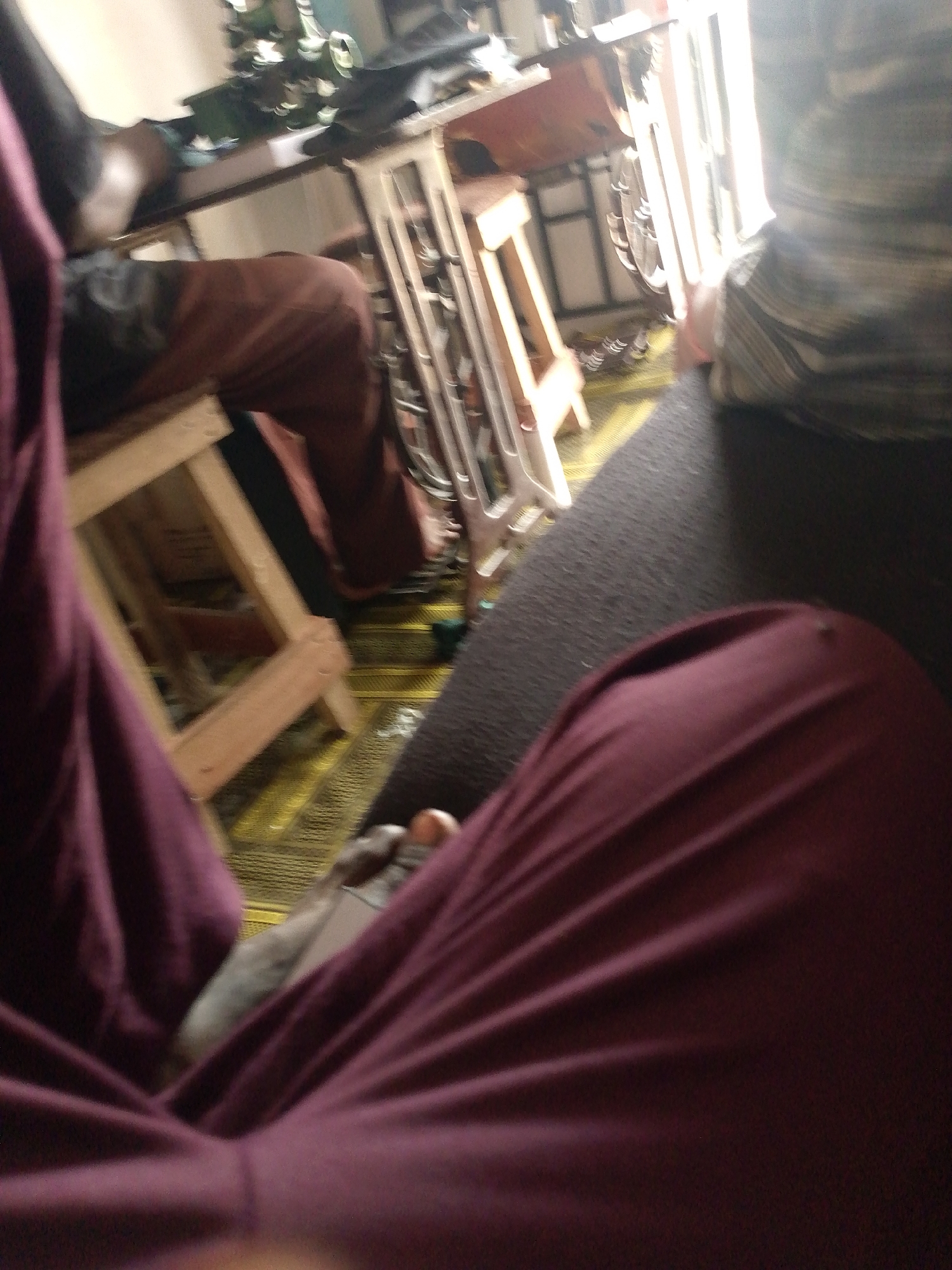 The image shows a close-up, low-angle perspective of a person's legs and torso, clad in dark burgundy fabric. The fabric has distinct folds and creases, suggesting it is loose-fitting clothing such as pants or a skirt. The person appears to be seated on a dark, textured surface, possibly a couch or carpet.
In the background, the scene is blurred but suggests an indoor environment. To the left, a wooden stool with a simple, angular design is visible, and behind it, a person's legs in reddish-brown trousers are also partially visible. Further back, there are hints of furniture and possibly other objects on a shelf or table, including metallic elements that might be part of a structure or appliance. A patterned rug with yellow and green accents covers the floor. The lighting suggests a brightly lit area, possibly near a window, with a bright light source in the upper right.
The overall impression is an informal, personal snapshot taken from a seated position, focusing on the lower body and immediate surroundings. There is no visible indication of the specific location in Jauro Yinu, Nigeria, as the image lacks any identifiable landmarks or context specific to that region.