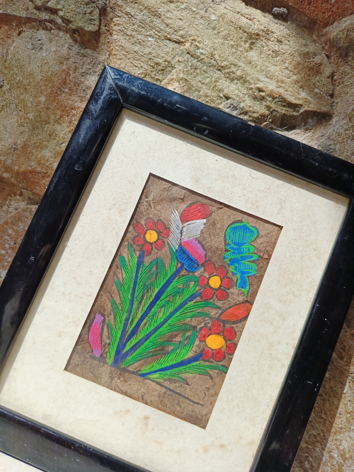 A framed piece of artwork rests against a textured, tan-colored stone surface. The frame is black, with some visible wear and possible dust. The artwork itself features a collection of brightly colored flowers and other abstract shapes. The colors are vibrant, including shades of red, orange, blue, green, and pink. The artwork is on a brown, possibly textured background. The photograph appears to have been taken indoors, and the lighting is somewhat uneven. The location is likely Los Robles, Venezuela, but the image does not provide any specific location clues within the city.