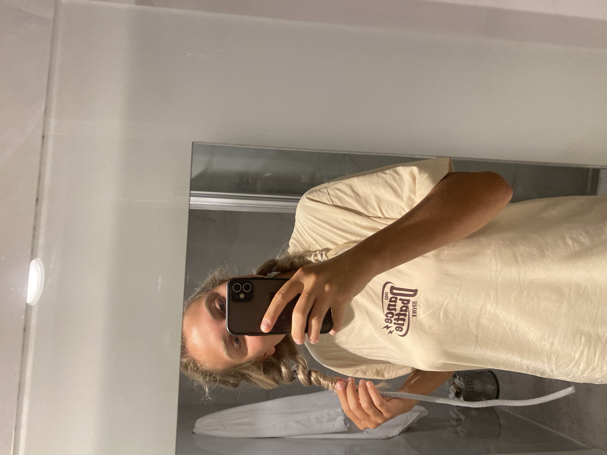 A young woman with braided blonde hair is taking a selfie in a bathroom, holding up her phone with her right hand. She is wearing a light-colored t-shirt with a graphic design. Her left hand is touching one of her braids. Behind her, a white bathrobe is hanging on a hook, and a silver trash can is visible. The bathroom appears to be modern, with a large mirror and tiled walls. There are no discernible features in the image that would indicate a location in Aksu, Turkey.