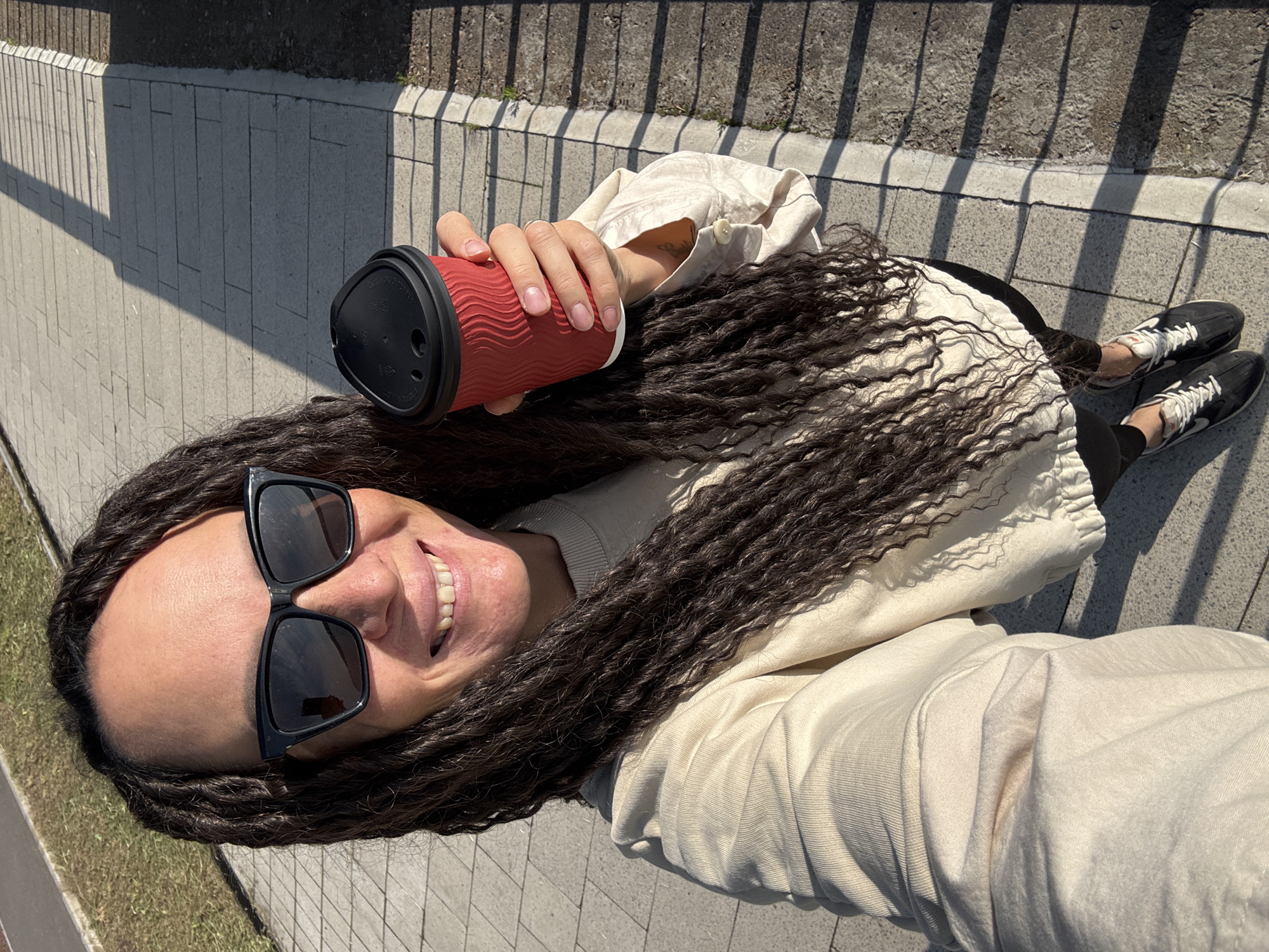 A high-angle selfie captures a person with long, dark dreadlocks lying or reclining outdoors in Chaikovsky, Russia, on a bright, sunny day. The individual is smiling broadly, revealing teeth, and wears large black square-framed sunglasses. They are dressed in a light-colored, possibly beige or cream-colored, top or jacket with long sleeves.
In their right hand, they hold a red, textured coffee cup with a black plastic lid. The lid has a small rectangular opening and two round holes. Visible on the lid is embossed text, which is partially legible as "CAUTION: CONTENTS..." and possibly a temperature warning or brand information. The left arm of the person is visible near the cup.
Their body and legs are partially visible, clad in what appears to be dark leggings or trousers, and black and white sneakers. The background consists of a paved walkway or ground made of rectangular grey pavers, with strong, elongated shadows cast by a railing or fence. The bright sunlight suggests a clear sky and daytime. The overall mood is cheerful and relaxed.