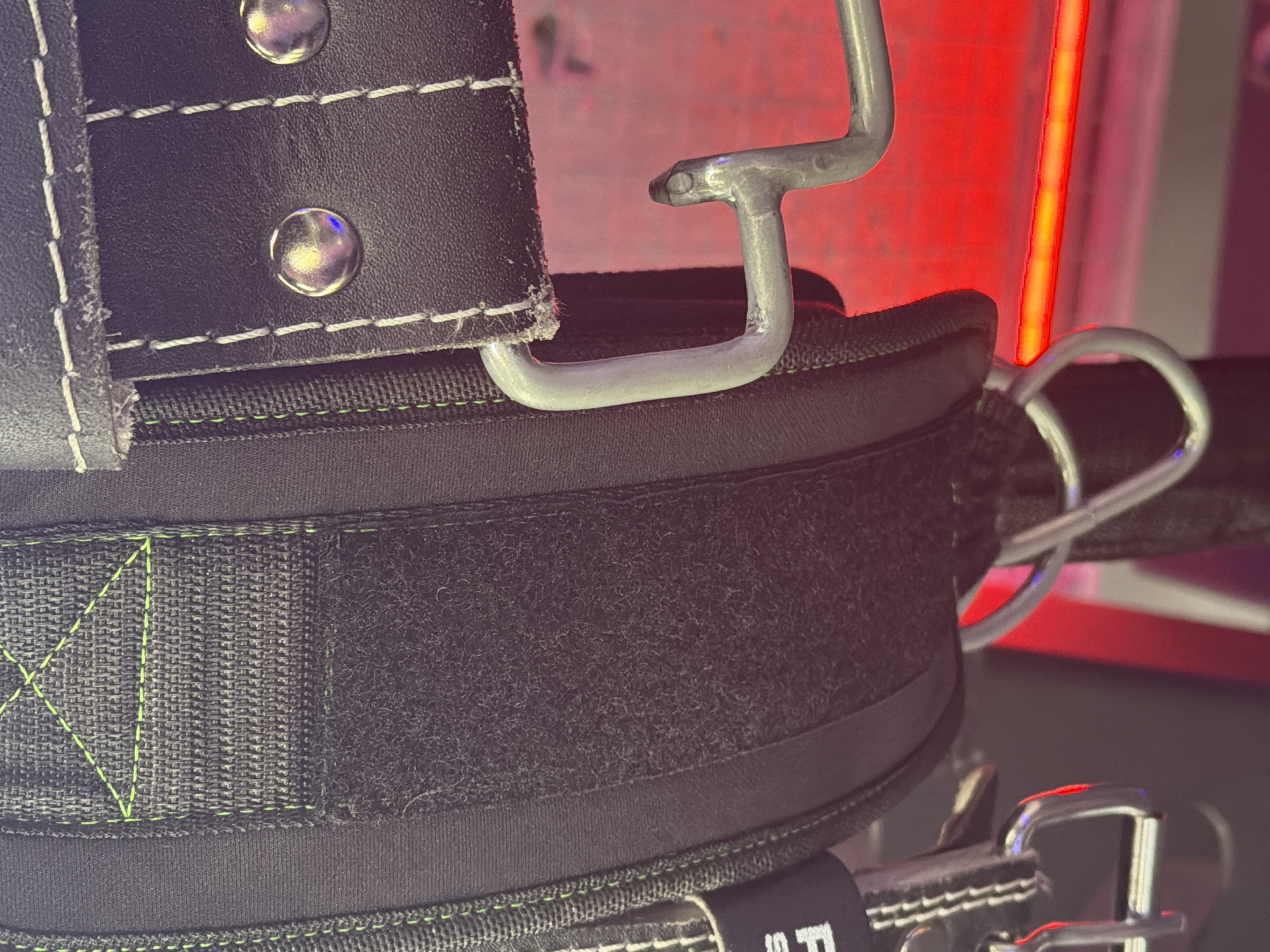 A close-up, low-angle, vertical, outdoor shot captures a black weightlifting belt with green stitching and chrome hardware. The belt features a velcro strap and a hook mechanism. The background is blurred and consists of a red light source. The image does not contain any discernible location information within the city of Unknown, Unknown, nor does it show any people or actions.