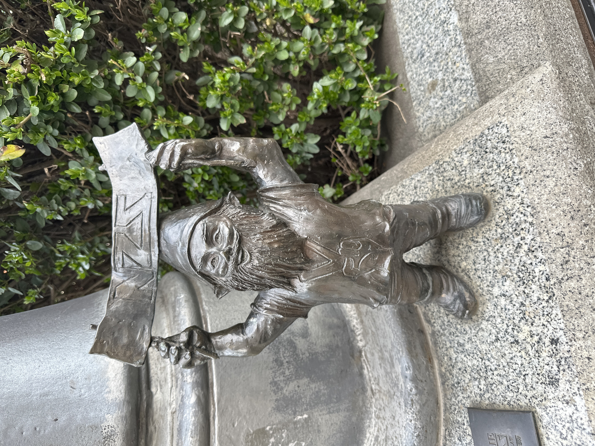 A bronze sculpture of a gnome, specifically one of the "Wrocław Dwarfs," is positioned outdoors in the city of Wrocław, Poland. The gnome is depicted with a long beard and a cap. It is holding a metallic scroll or banner featuring vertically stacked symbols. The sculpture is placed on a grey, stone-like surface, likely a part of the urban infrastructure. Behind the gnome is a cluster of green bushes or shrubs. A small plaque with Japanese characters is visible on the stone surface near the bottom right.