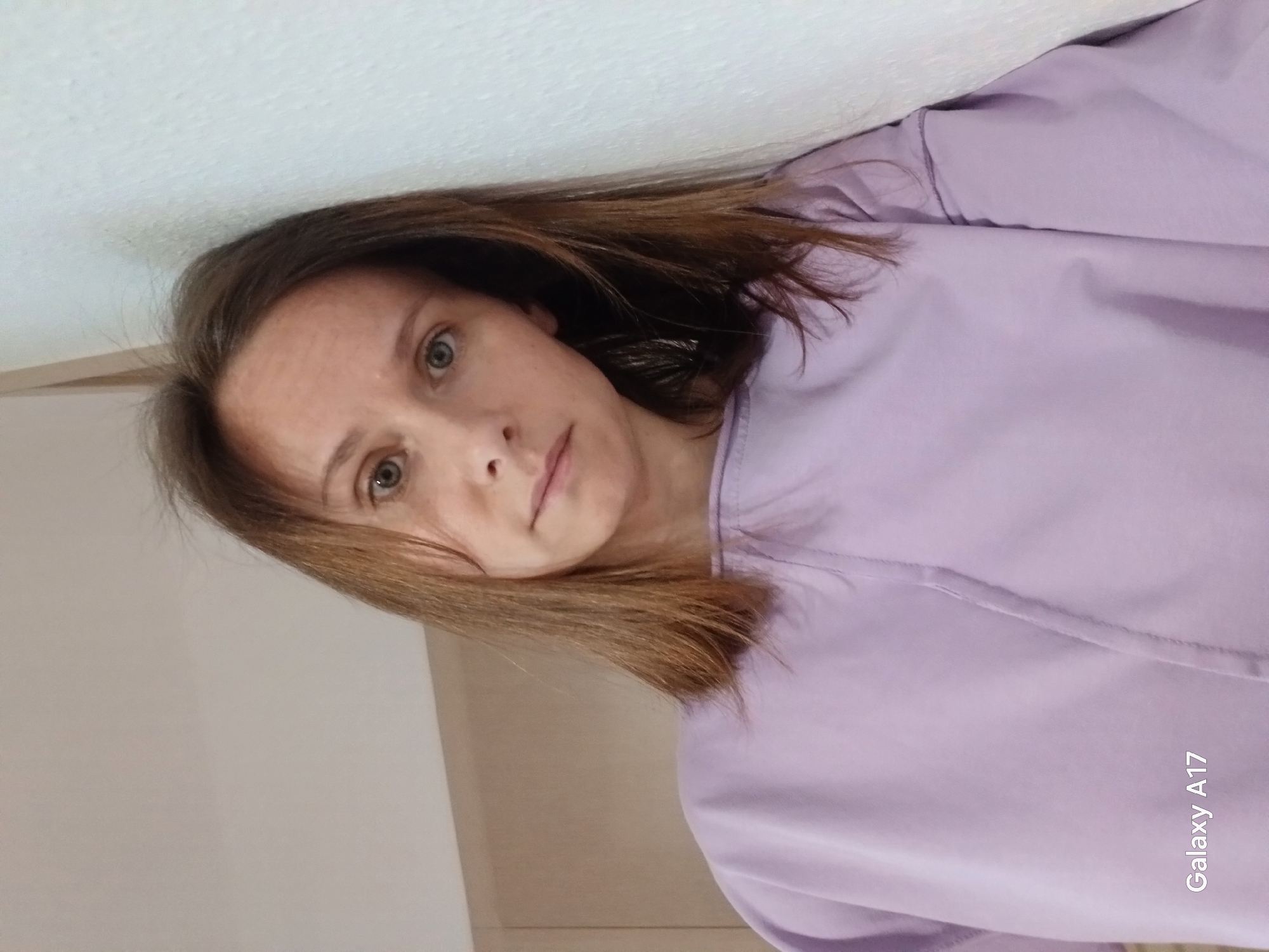 A woman with shoulder-length brown hair and light eyes is seen indoors, wearing a lilac top. She is leaning against a white wall. The location appears to be a room with light-colored walls and minimal furnishings. The city of Yekaterinburg, Russia, is not discernable from the image. There are no visible actions or interactions taking place. The environment appears to be a simple, indoor setting. The bottom right corner of the image displays "Galaxy A17".