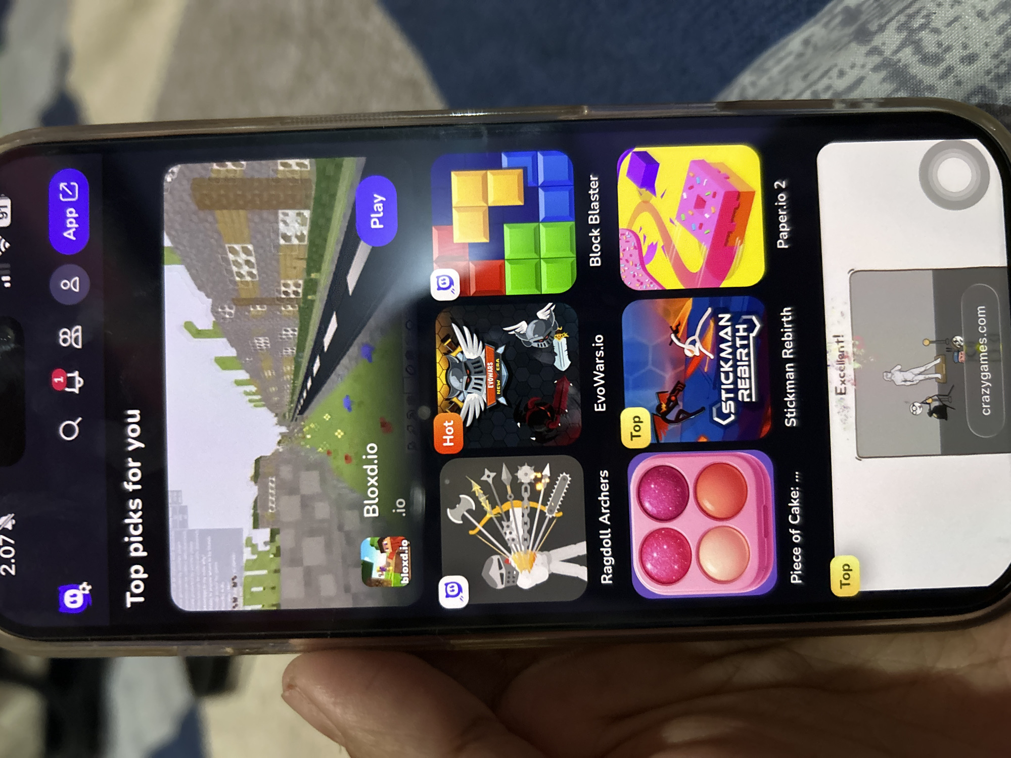 The image shows a smartphone screen displaying a mobile application with various game recommendations. The user is holding the phone, and a portion of their hand is visible on the left side. The application interface features a "Top picks for you" section, showcasing several game icons and titles, including "Bloxd.io," "Ragdoll Archers," "EvoWars.io," "Block Blaster," "Piece of Cake: ...," "Stickman Rebirth," and "Paper.io 2." Some games have "Hot" or "Top" labels indicating their popularity. At the top of the screen, there is a status bar showing the time "2.07," battery indicator "91," and network signal. There are also navigation icons for search, notifications, and user profiles. The background of the phone screen displays a rendered 3D environment resembling a cityscape or a game level. The overall scene is indoors, and the lighting suggests a normal ambient environment. There is no visible indication that this scene is located in Depok, Indonesia.