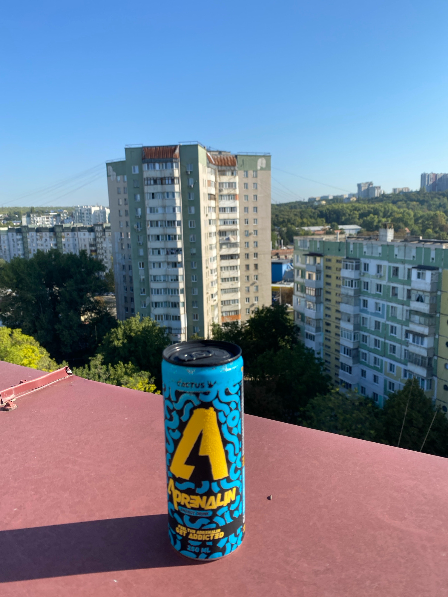 An eye-level, medium shot captures a bright blue can of "ADRENALIN" energy drink positioned on a rusty red, metallic surface, possibly a rooftop. The can is adorned with a large yellow "A" symbol and a wave-like pattern in black. The background reveals a clear, azure sky above a cityscape characterized by numerous multi-story apartment buildings. Some buildings exhibit faded pastel facades in green, yellow, and white, with visible balconies and numerous windows. Lush green trees are interspersed between the buildings and in the foreground, suggesting a temperate climate. The overall scene suggests an outdoor urban setting on a sunny day. The image is consistent with a view from a building in Chișinău, Moldova, given the architectural style of the apartment blocks. There are no people or animals visible in the frame.