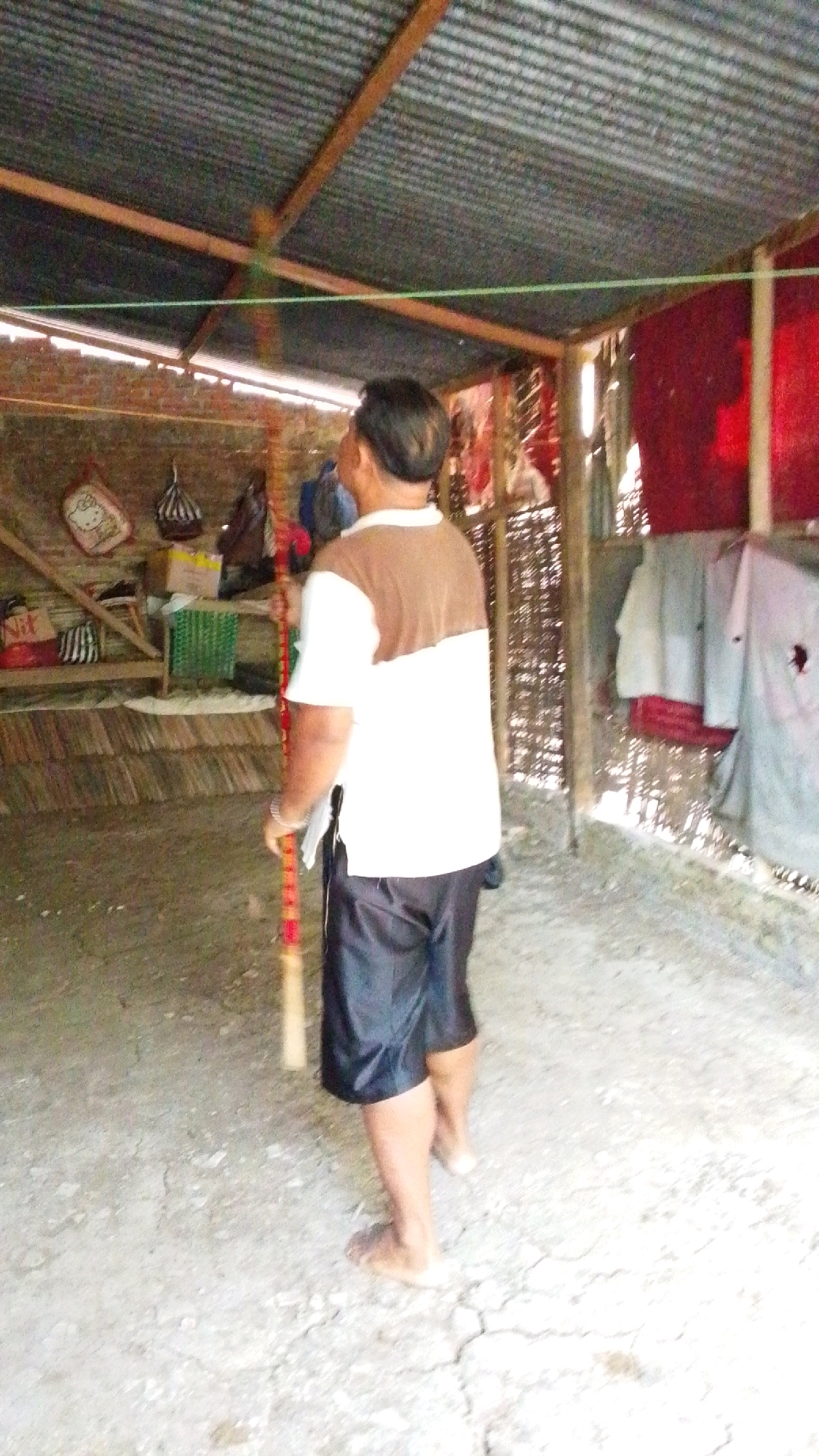 A man is standing in what appears to be a covered outdoor area. He is facing away from the camera, wearing a white t-shirt with a brown section across the chest and shoulders, and black shorts. He is holding a long, thin object, possibly a stick or a pole, which has red and yellow bands on it. The ground is a mix of dirt and gravel. In the background, there is a corrugated metal roof supported by wooden beams. To the left, there are shelves with various items, including bags and what looks like a box. To the right, there is a woven bamboo-like wall and some clothes hanging. The environment suggests a rustic or informal setting. It is not possible to determine the specific location within Lawu, Indonesia from the provided image.