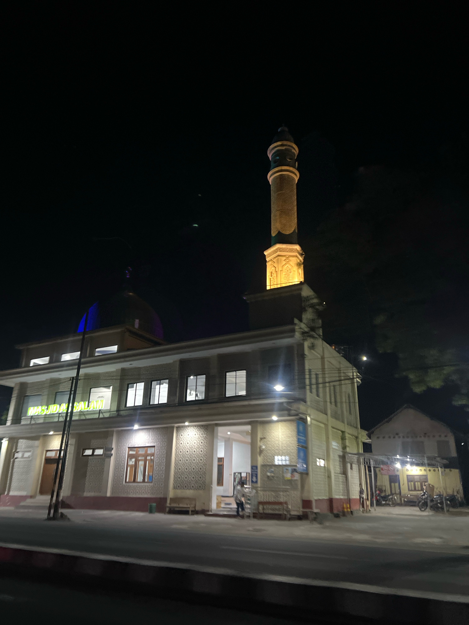 This is a nighttime, low-angle, full shot of a mosque in Jumoyo, Indonesia. The mosque has a prominent minaret illuminated with yellow light and a blue dome. The building is lit with white lights, revealing the "MASJID BALAM" sign above the entrance. The mosque is situated next to a street, with a few motorcycles parked in front of a separate building in the background. The scene is dark, with the mosque and surrounding structures being the main illuminated subjects.