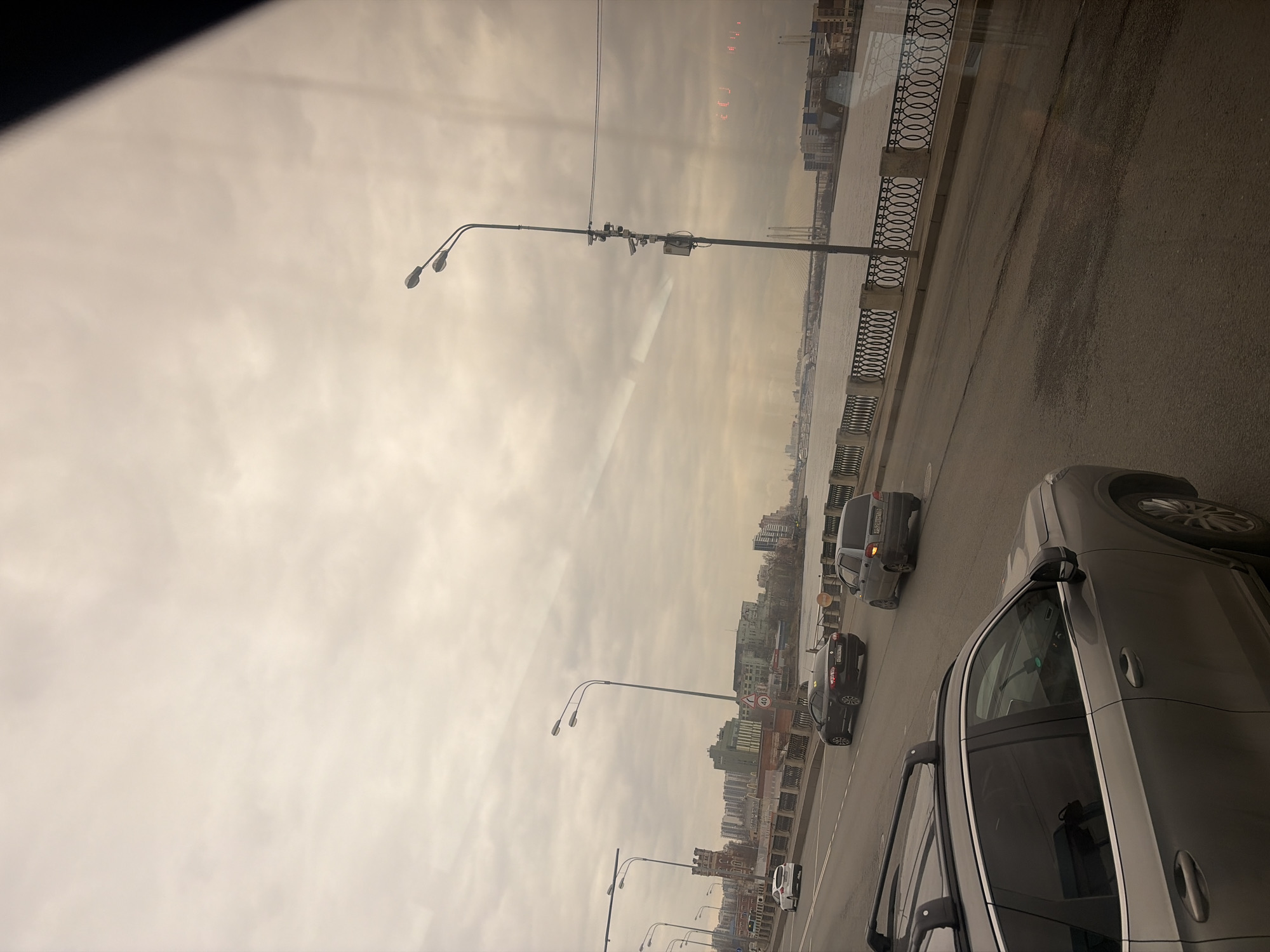 The image shows a view from a vehicle traveling on a bridge in Saint Petersburg, Russia. The shot captures a gray, overcast sky and a segment of the river Neva alongside the bridge. The right side of the frame includes the asphalt of the bridge, part of the vehicle from which the photo was taken, and a decorative metal railing that lines the riverbank. Traffic can be seen moving along the bridge, with several cars ahead of the vehicle from which the photo was taken. Street lights line the bridge, and buildings can be seen along the riverbank in the distance. The atmosphere appears to be hazy or smoggy.