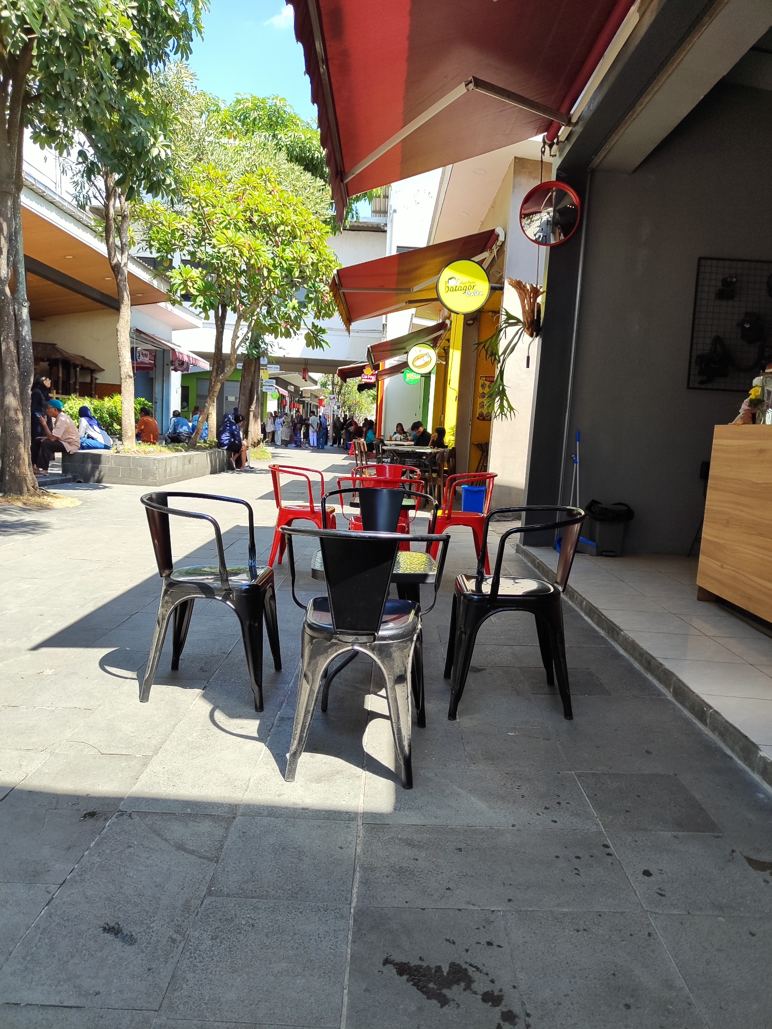 The image depicts an outdoor seating area for a cafe or restaurant on a sunny day. In the foreground, there are four black metal chairs arranged around a small, dark-colored table. Behind these, a red metal chair is visible. The seating area is paved with large, grey rectangular tiles, with strong shadows cast by the chairs and overhead structures.

In the midground, there is a pathway with more trees and people seated on a low wall or bench to the left, appearing to be patrons of the establishments lining the walkway. Further back, more tables and chairs are visible, along with the facades of shops and cafes, some with red awnings. Signs are visible, including a yellow circular sign with the text "Batagor by..." and a smaller green sign with a depiction of a food item.

The background shows more of the walkway, with additional people browsing or dining. The environment appears to be a pedestrian zone within a commercial area, possibly a market or a shopping street. The buildings have a modern architectural style. The scene suggests a lively, public space during daylight hours in Sempusari, Indonesia.