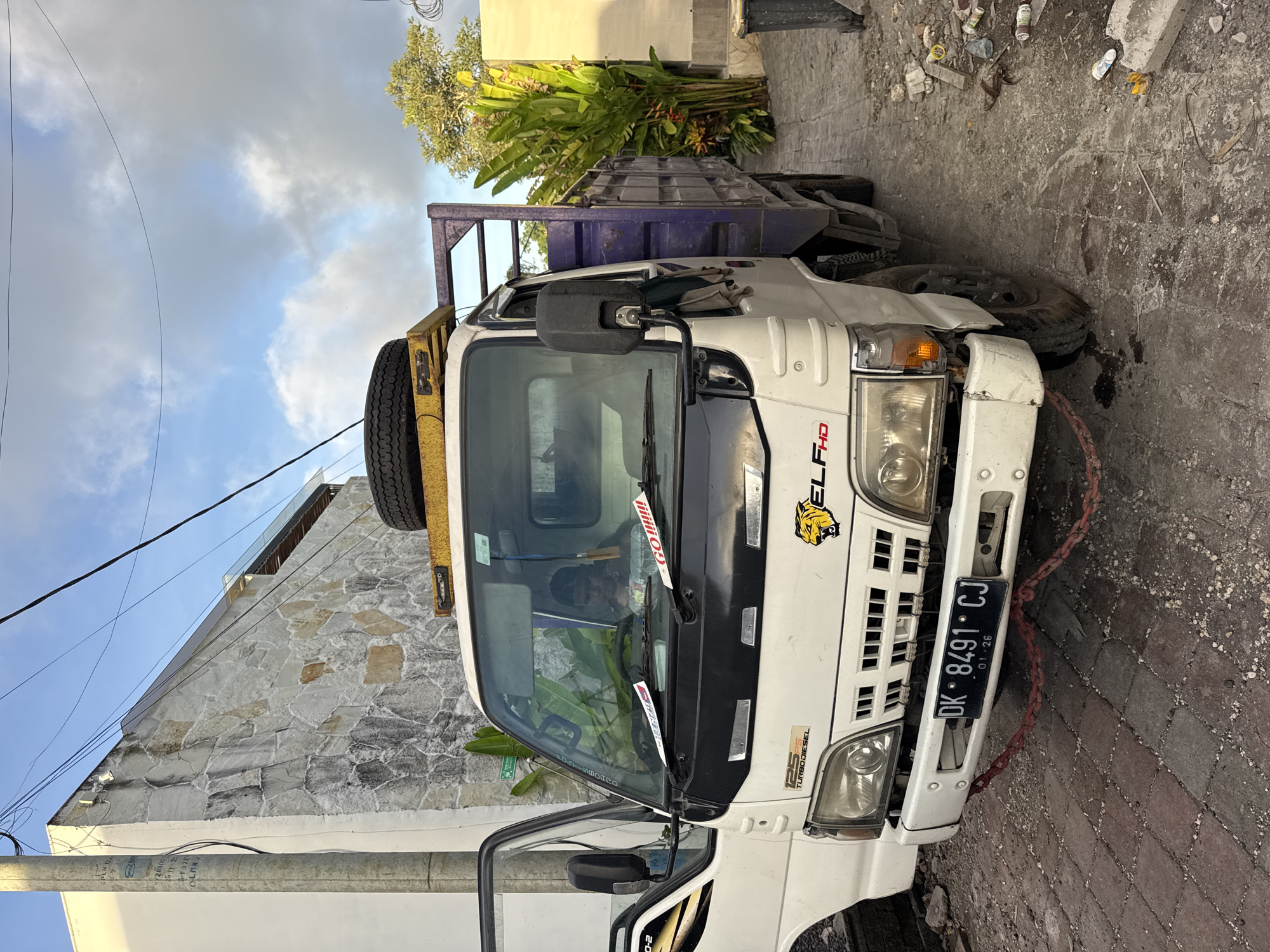 A medium shot of a white Isuzu Elf HD truck with a blue bed, parked on a brick-paved surface in Kuta Selatan, Bali, Indonesia. The truck has a yellow spare tire mounted above the cab. The license plate reads "DK 8491 CJ" with an expiration date of 01-26. The truck's front is facing the viewer's lower left.
The truck is parked in a narrow alley next to a building with a rough stone facade. A utility pole with wires is visible in the upper left. The ground around the truck shows signs of debris and trash. A cluster of plants is located next to the truck's bed.