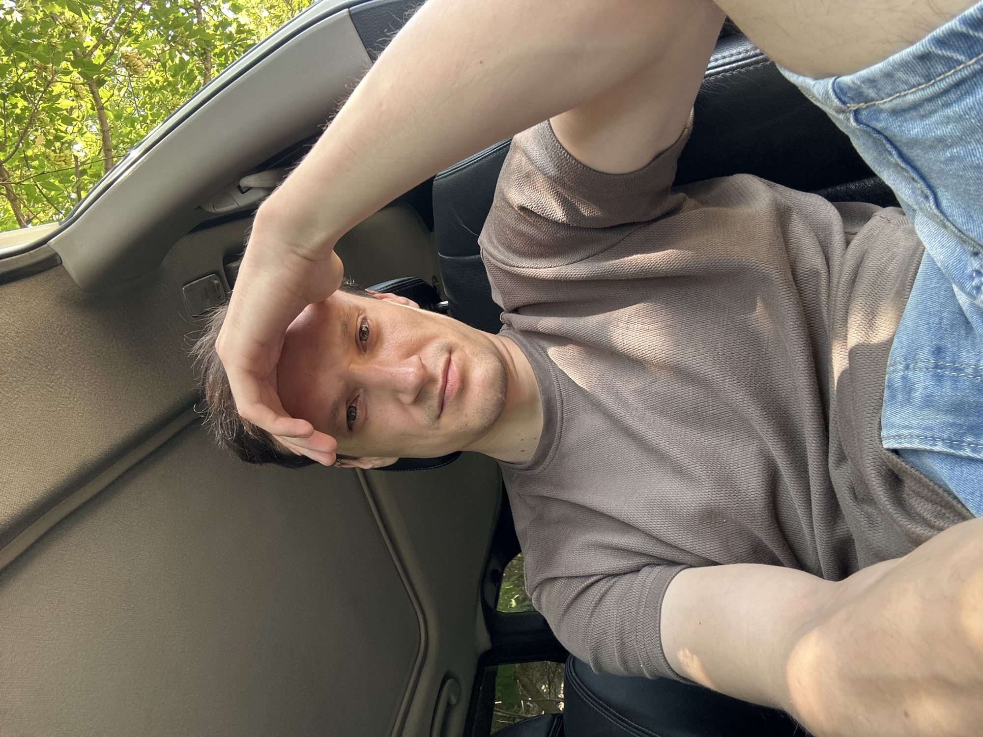 A man is sitting in the driver's seat of a car, his right hand resting on his forehead. He is looking directly at the camera. The interior of the car is visible, including the ceiling with a sunroof and dark leather seats. Outside the car, trees with green foliage are visible, suggesting an outdoor, possibly rural or suburban setting. The man is wearing a brown t-shirt and blue denim shorts. The image appears to be a selfie taken inside the vehicle. No specific location in Makiv, Ukraine is evident.