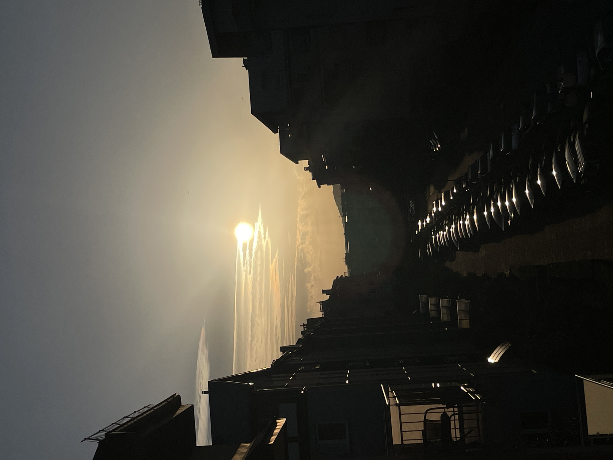 The image depicts a street scene in an urban environment, likely during sunset. Tall, silhouetted buildings line both sides of a narrow street. The sun is visible in the upper center of the frame, partially obscured by clouds and casting a warm glow across the sky. A long row of parked cars, their surfaces reflecting the sunlight, fills the street. The overall impression is one of a quiet urban evening, with the setting sun creating dramatic shadows and highlights. The scene appears to be in a residential area, with balconies and windows visible on the buildings.