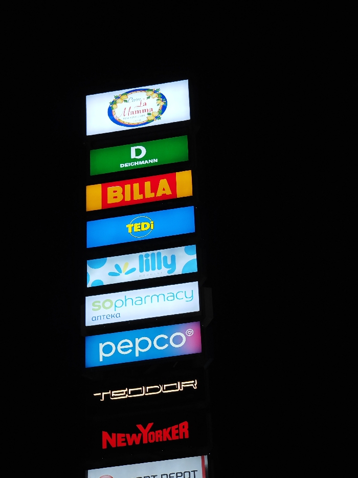 The image displays a vertically oriented, illuminated multi-tenant pylon sign against a dark background, indicating nighttime conditions. This sign is located at Retail Park Shumen, Kaliakra Street 1, 9700 Shumen, Bulgaria. The structure consists of stacked rectangular lightboxes and backlit signs, each featuring the logo and name of a commercial establishment. From top to bottom, the visible businesses are: Osteria "La Mamma", DEICHMANN, BILLA, TEDi, Lilly Drogerie, SoPharmacy, PEPCO, TEODOR, NEW YORKER, and the upper portion of SPORT DEPOT. All displayed logos are brightly illuminated. No individuals or additional environmental details are visible.