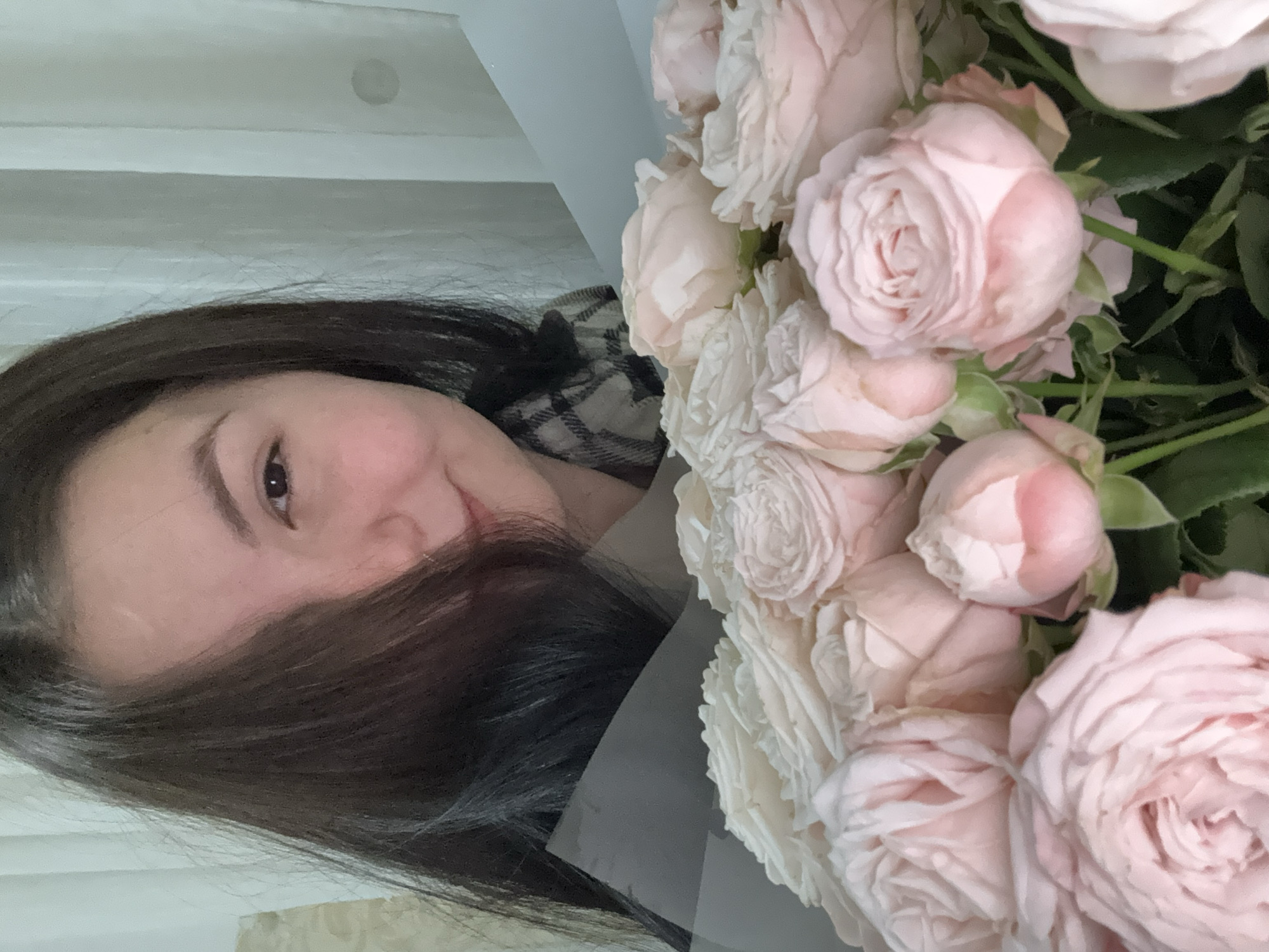 The image shows a woman with dark hair holding a bouquet of pale pink roses. Her face is partially obscured by her hair. She is wearing a checkered scarf or top. The background appears to be a light-colored interior. There is no information in the image to suggest it was taken in the city of Фоки, Russia.