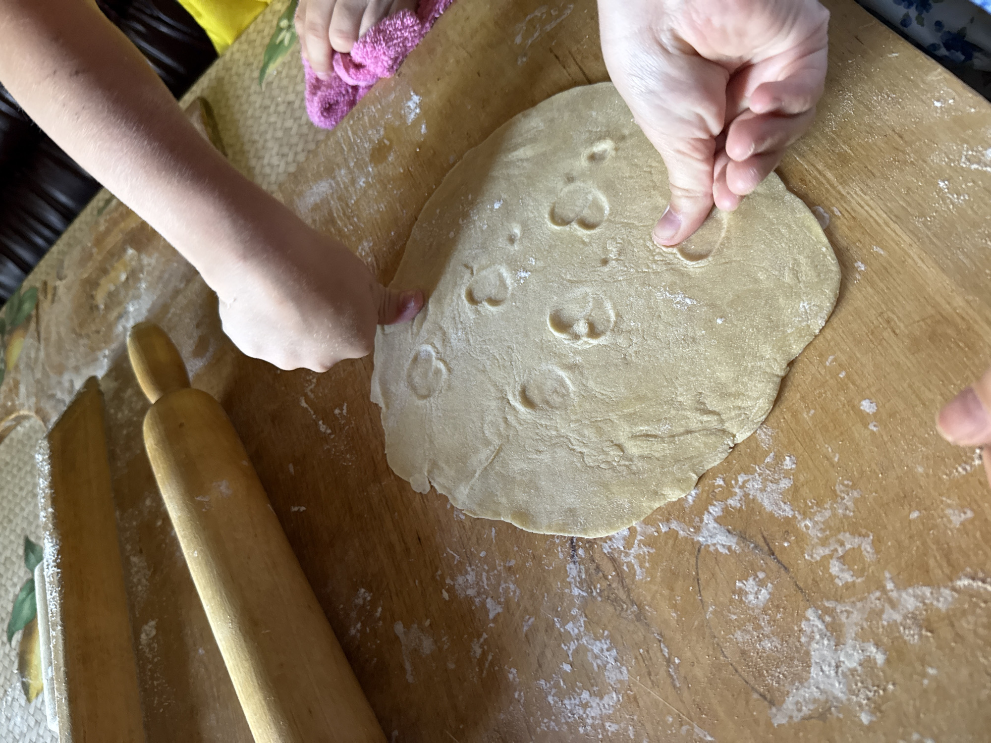 This image depicts a close-up overhead view of two pairs of hands interacting with dough on a wooden surface. The scene is not located in Valday, Russia. One person's hands are gently pressing indentations into a large, circular piece of light-colored dough that is spread across the wooden table. These indentations appear to be in the shape of small hearts. Another person's hands are visible on the right side, holding a small, pink, textured cloth. A wooden rolling pin lies horizontally in the upper left quadrant of the image, and another similar object is partially visible behind it. The wooden surface is dusted with flour, indicating food preparation. The lighting is warm and natural, casting soft shadows. The background is out of focus but suggests an indoor setting with dark furniture.