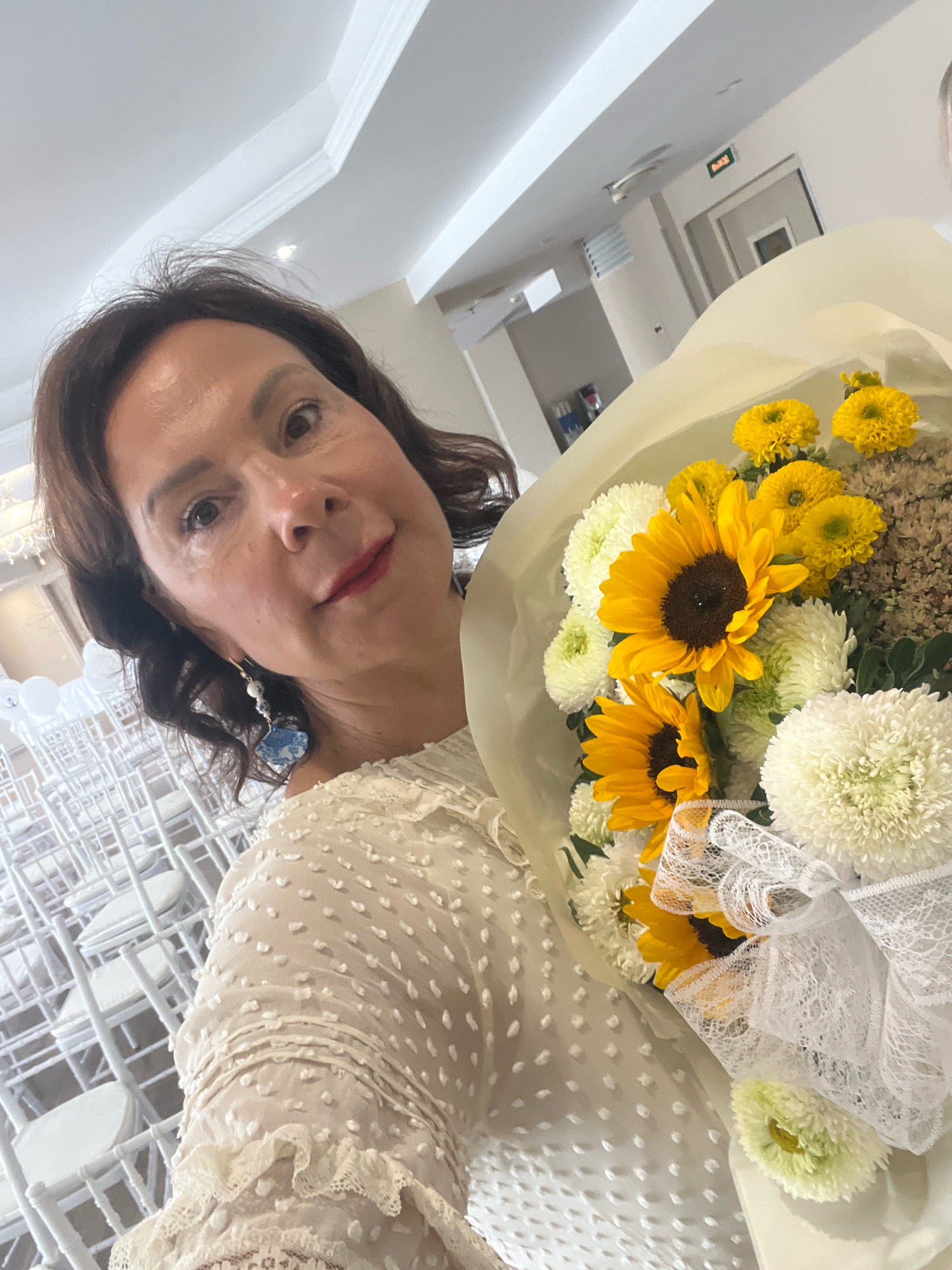 The image shows a medium shot of a woman of European descent, with shoulder-length dark brown hair, holding a bouquet of flowers. She is wearing a white, textured dress with lace trim. The bouquet contains sunflowers, white chrysanthemums, and smaller yellow flowers. In the background, rows of empty white chairs are visible, suggesting an event space like a banquet hall or wedding venue. A sign with Cyrillic text indicating "EXIT" is visible in the upper right background, suggesting the location is in Russia. The lighting is bright and appears to be from overhead sources. The woman is looking towards the camera, and her expression is neutral. The setting is indoors, with a ceiling and walls visible.