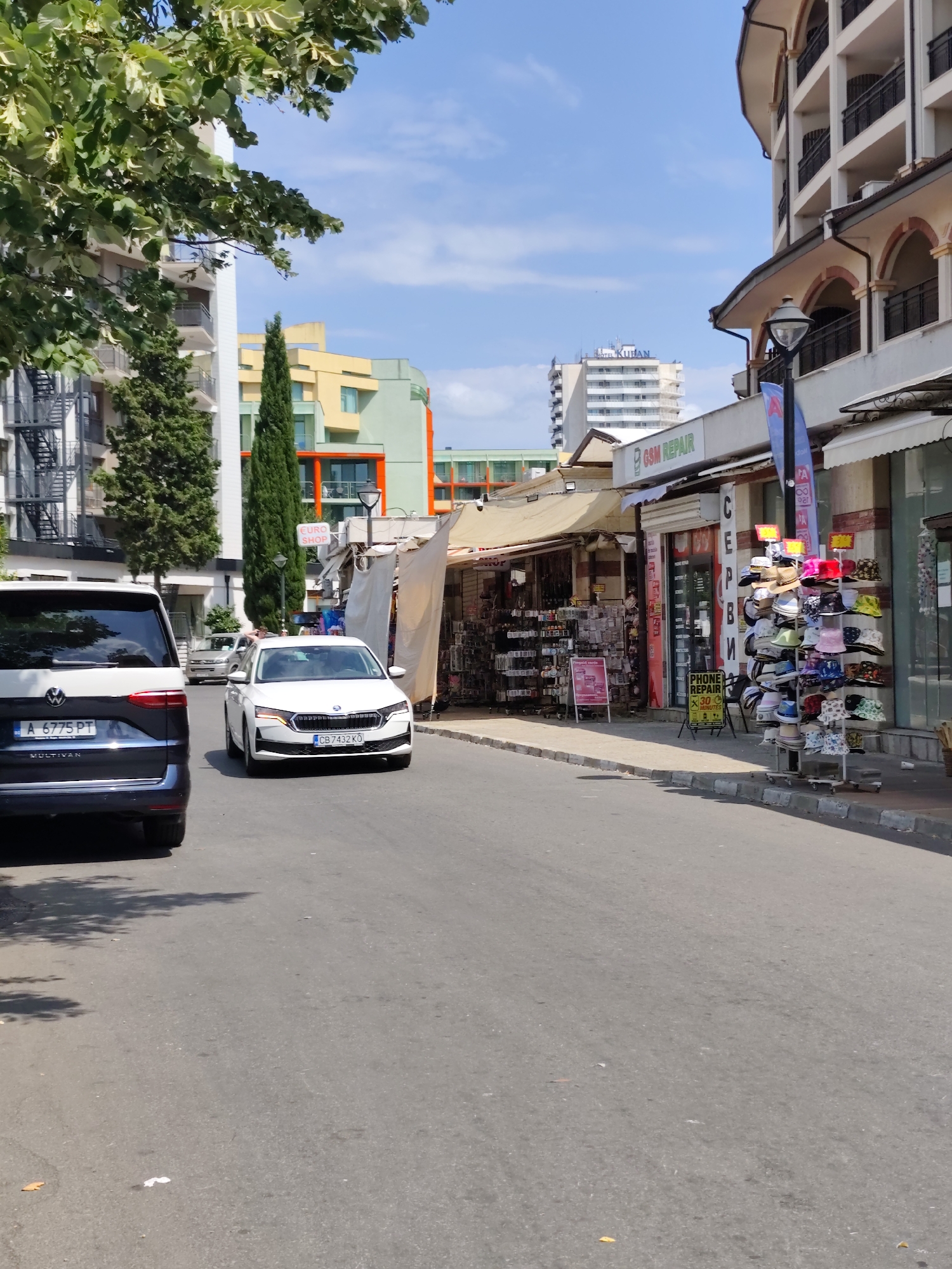 In Nesebar, Bulgaria, a street scene unfolds under a bright, partly cloudy sky. The camera, positioned low and looking down the street, captures a sun-drenched asphalt road. On the left, a dark blue and white van, a Volkswagen Multivan with a Bulgarian license plate "A 6775 PT", is parked on the side of the road, its rear end prominently featured. Further down the road, a white Skoda sedan with a Bulgarian license plate "CB 7432 KO" drives away from the camera.
The street is lined with buildings. On the right, a multi-story building with arched balconies and beige facade rises, featuring shops at ground level. One shop displays hats on a rack, showcasing various styles and colors. Another shop has a sign that reads "GSM REPAIR" and "PHONE REPAIR 30 MINUTES". A tall, slender cypress tree stands between the parked van and a colorful building with orange and yellow elements. Beyond that, more buildings are visible, with a distant tall, modern hotel building with the name "HOTEL KUBAN" visible at the top. The environment appears to be a commercial area, possibly a tourist destination, with shops and a main road. Shadows from trees are cast on the road, indicating the sunny weather.