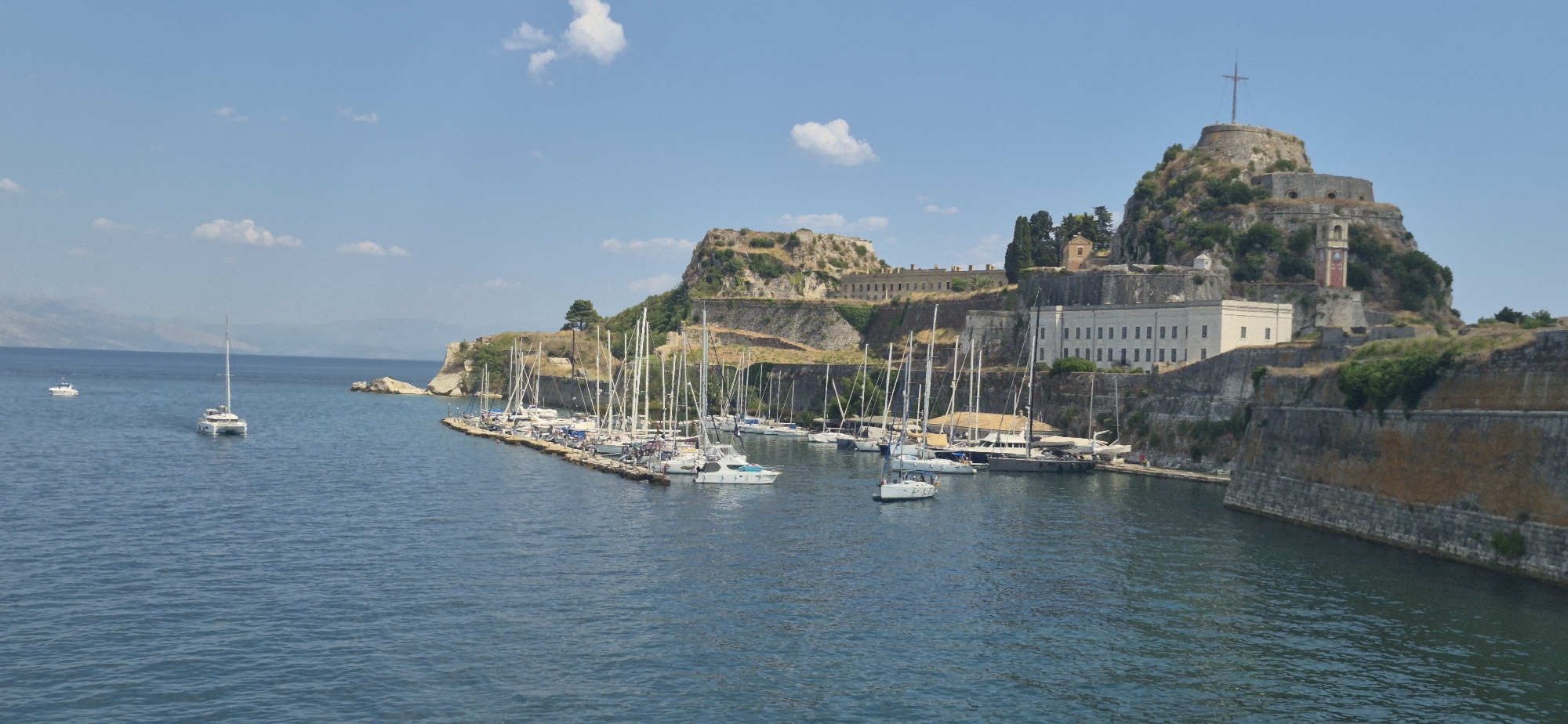 The image depicts a picturesque harbor in Corfu, Greece, likely the Old Fortress marina, showcasing a serene blue body of water dotted with numerous white sailboats and a few other vessels. The boats are docked along a long pier, their masts reaching towards a clear blue sky with a few scattered white clouds. In the background, a formidable ancient fortress, with its weathered stone walls and fortifications, rises majestically from a rocky outcrop. The fortress complex includes several buildings, one of which is a light-colored structure with many windows, and a distinctive clock tower with a red brick base. The overall scene is bathed in bright sunlight, suggesting a pleasant day in this historic coastal city.