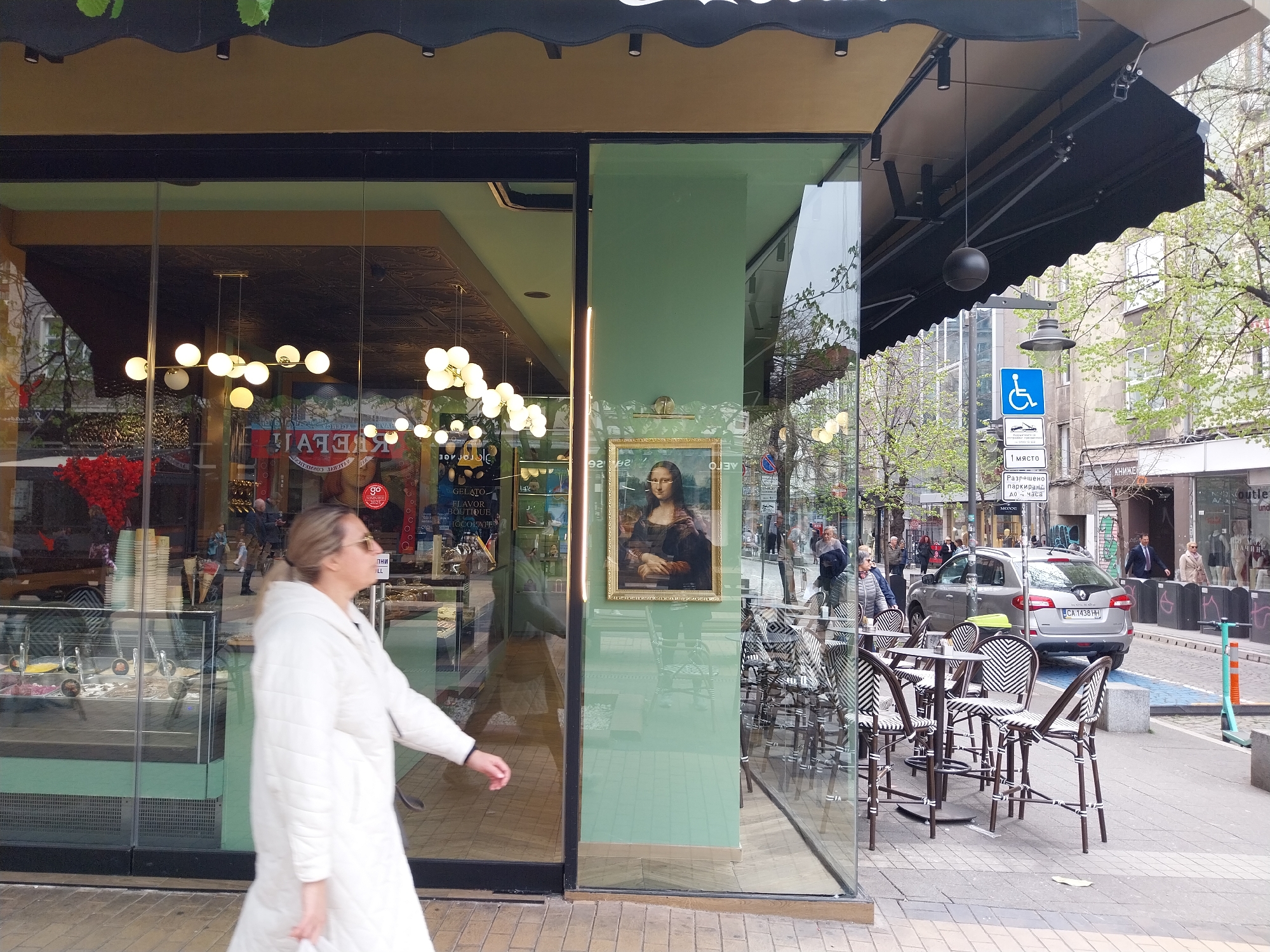 Here is a detailed description of the image:
The image captures a street scene in an urban area with a building featuring a storefront. A woman in a white coat is walking from left to right in the foreground. The store is a "Gelato Flavor Boutique Chocolat" shop with "go" logo, as written on the window. Inside, there are displays of gelato flavors and decorations.
The storefront has a section that is green, and mounted on this is a framed picture of the Mona Lisa. A black awning covers the top part of the building.
Outside, there are tables and chairs set up for outdoor seating. Behind the seating area, there is a street with parked cars and pedestrians. A blue handicapped parking sign is visible on a pole.
Based on the license plate on a car ("CA 1438 HH") and the language on the parking sign, the location is likely in Bulgaria, although the city is unknown. The image is taken during daylight, with sunlight illuminating the street.