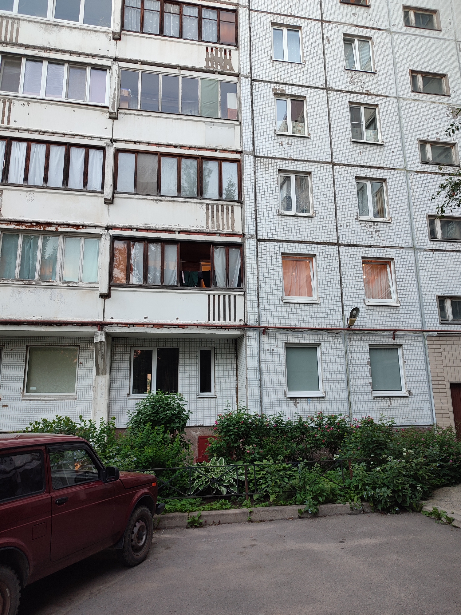 The image depicts a multi-story apartment building in Vsevolozhsk, Russia. The building has a light-colored tiled facade with numerous windows. Some windows are closed, while others appear to be open, revealing curtains or internal views. On the ground level, there is a patch of green vegetation with bushes and some flowering plants, bordered by a metal fence. A dark red, three-door SUV, possibly a Lada Niva, is parked on the asphalt in front of the building. The overall scene suggests a residential area within the city.