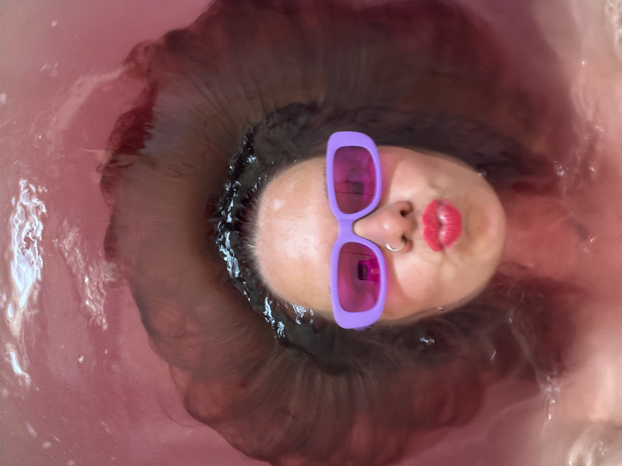 A person lies in pink-tinted water, possibly a bathtub, with their face partially submerged. They wear purple sunglasses and bright red lipstick. The surrounding water is also pink. Dark hair surrounds the person's face, and a silver nose ring is visible. The location is unknown.
