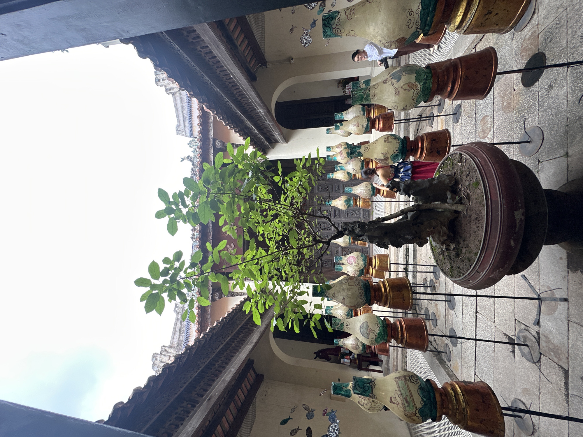 This image shows an interior courtyard with ornate architecture, likely a temple or historical building, located in Hanoi, Vietnam. The courtyard is paved with stone and features a central bonsai tree in a large pot. Surrounding the bonsai are numerous decorative ceramic vases or urns displayed on golden pedestals with thin metal stands. These vases are adorned with painted scenes and patterns. In the background, two women are visible. One, on the left, is seen from the back, wearing a colorful dress. The other, on the right, is facing towards the center of the courtyard, holding a camera and wearing a white shirt and dark pants. The architecture includes traditional tiled roofs and intricately carved wooden eaves. The sky above is bright and overcast.