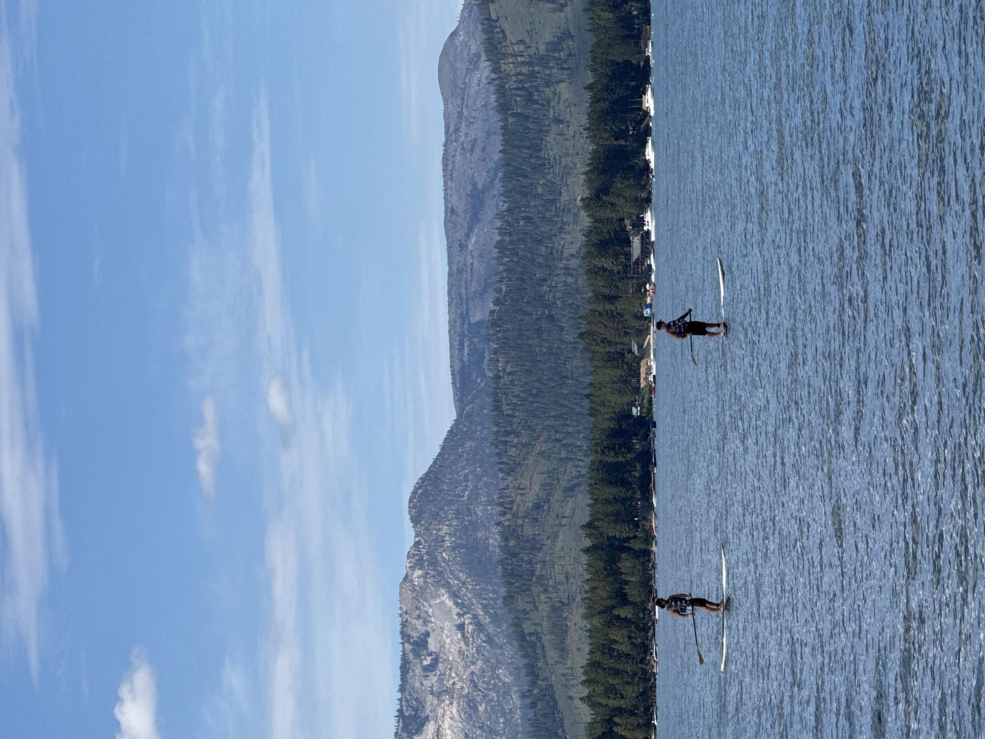Two individuals are paddleboarding on a calm, blue lake under a bright, partly cloudy sky. The person on the left is wearing a dark tank top and shorts, and the person on the right is wearing a dark tank top and striped shorts. Both are standing on white paddleboards and holding paddles. In the background, a dense forest of evergreen trees lines the shore, with a mountain range visible behind the trees. Several boats are moored along the shoreline. The scene appears to be set in a natural, recreational area, likely a popular tourist destination like Lake Tahoe, near Stateline, Nevada.