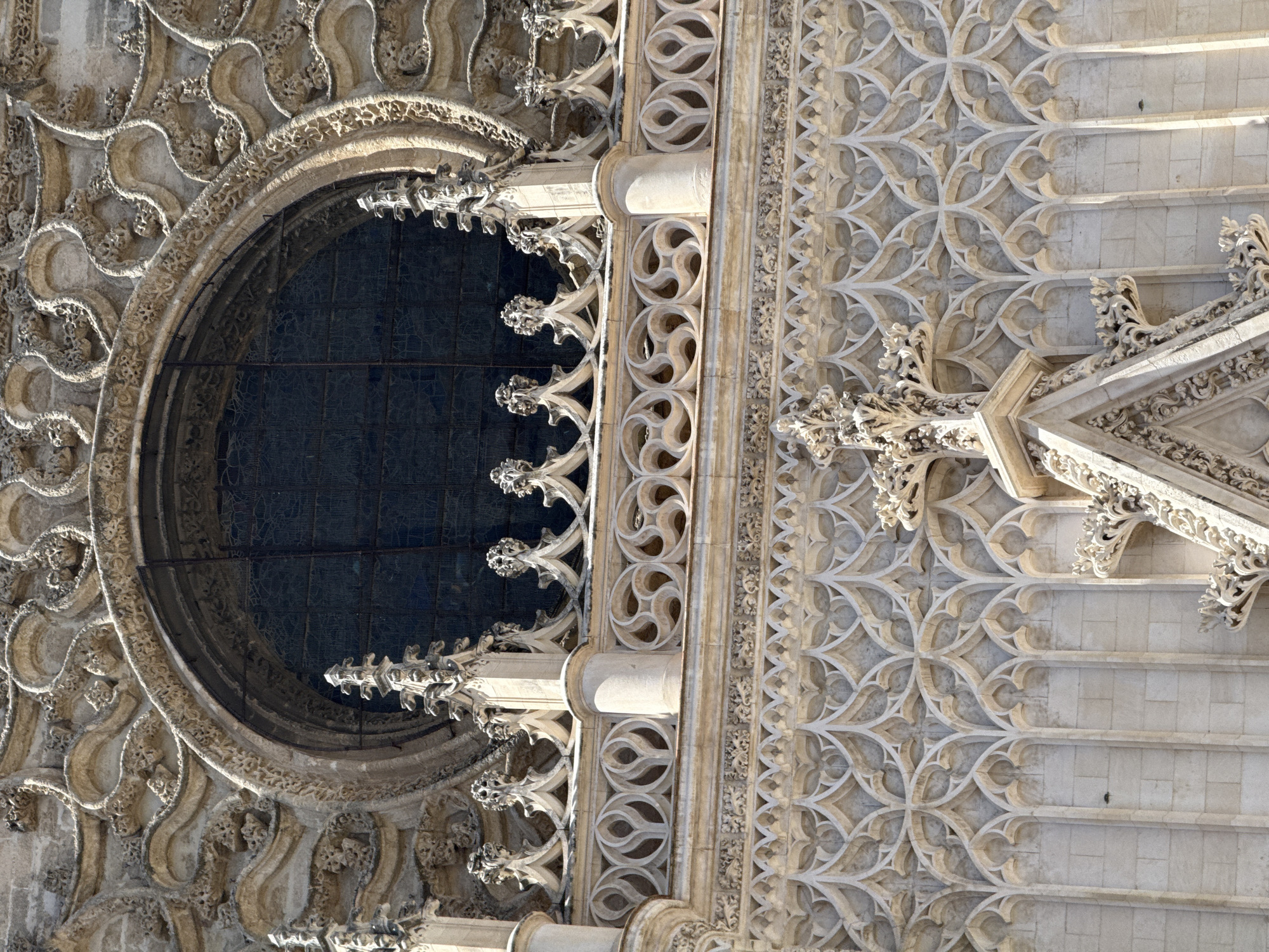 The image shows a detailed view of the exterior of the Seville Cathedral in Seville, Spain. The photograph focuses on the ornate Gothic architecture, particularly a large circular window (oculus) with intricate tracery. Below the window, a decorative frieze with repeating geometric patterns made of stone is visible. Further down, a section of the cathedral's facade displays delicate stone carvings and vertical elements that suggest buttresses or decorative pilasters. The material appears to be light-colored stone, weathered by time. The overall impression is one of grandeur and detailed craftsmanship. There are no people or any dynamic actions depicted in the scene.